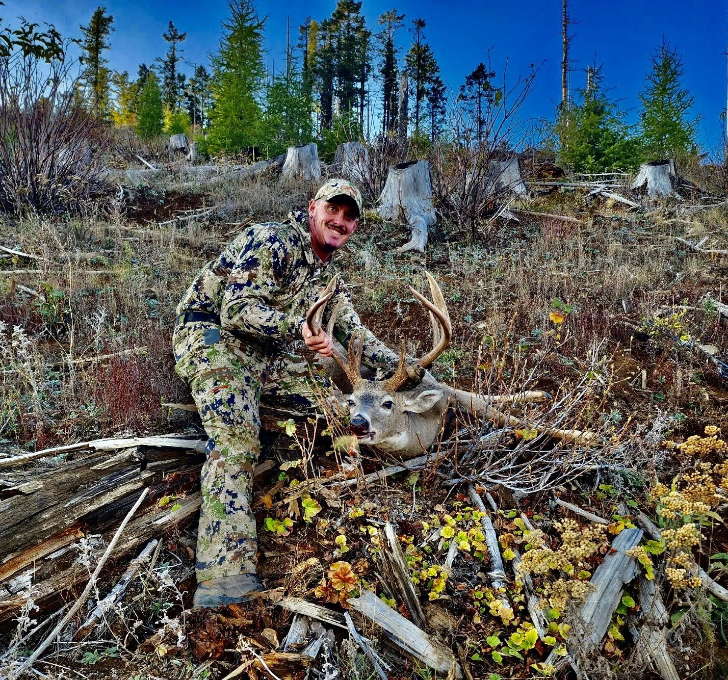 A man dressed in camouflage outdoor clothing is posing with a large deer with antlers in a forested area, surrounded by trees and fallen branches.