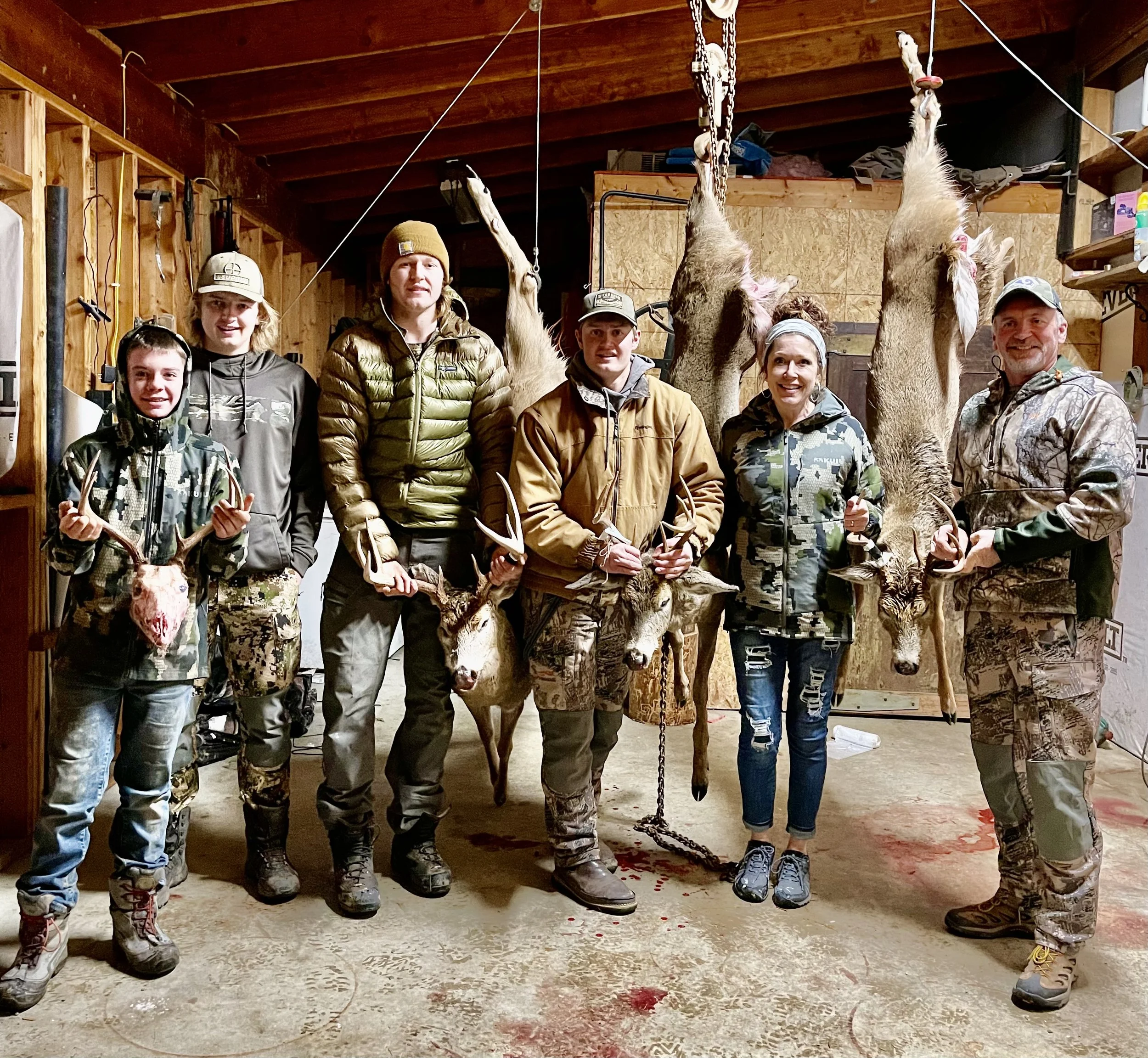 Group of six people in camo and outdoor clothing in a wooden hunting lodge, holding deer and elk antlers, with two hanging deer carcasses behind them and blood on the floor.