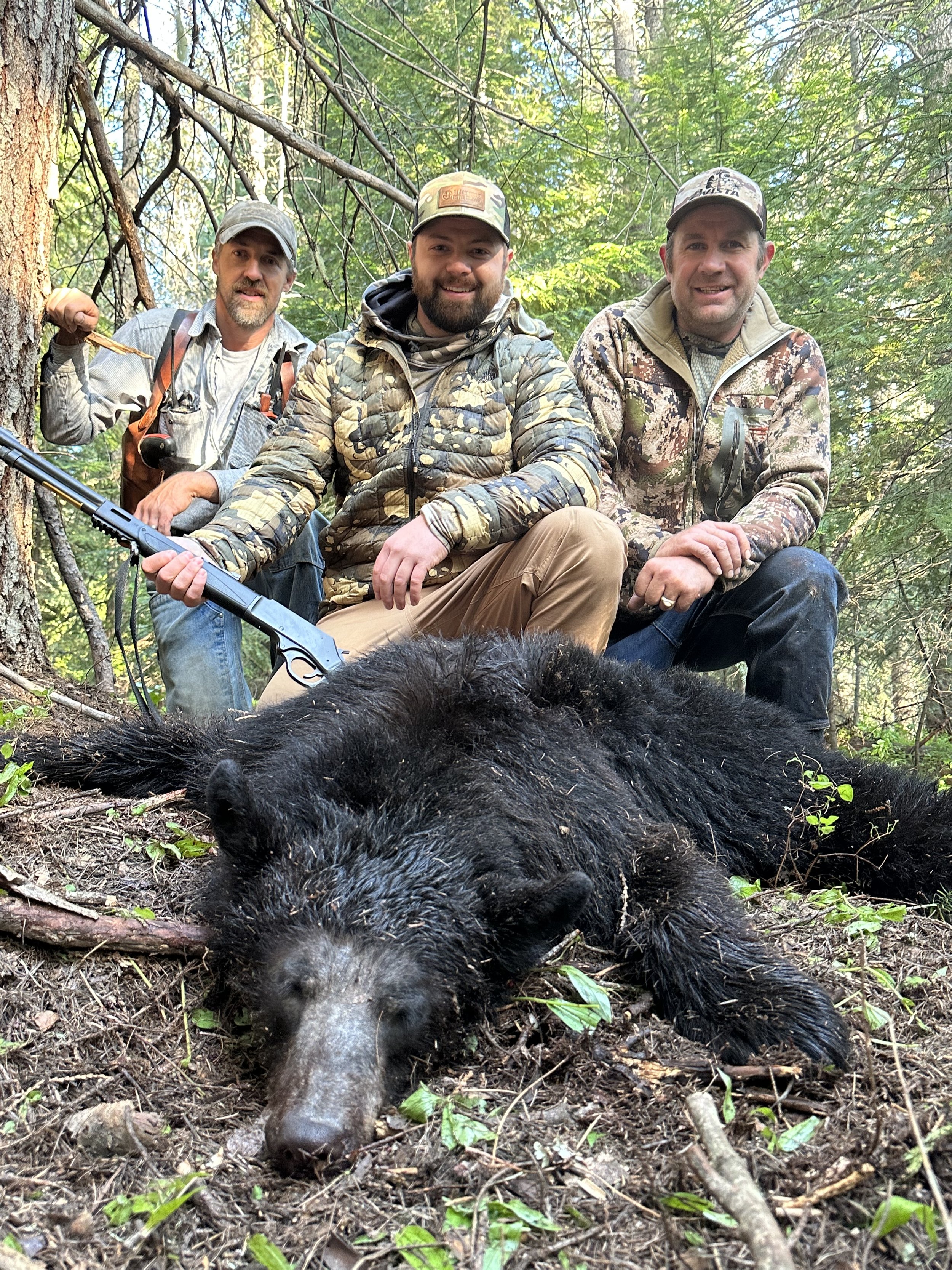 Three men in camouflage and outdoor clothing kneel in the forest behind a dead black bear lying on the ground, with green trees and branches overhead.