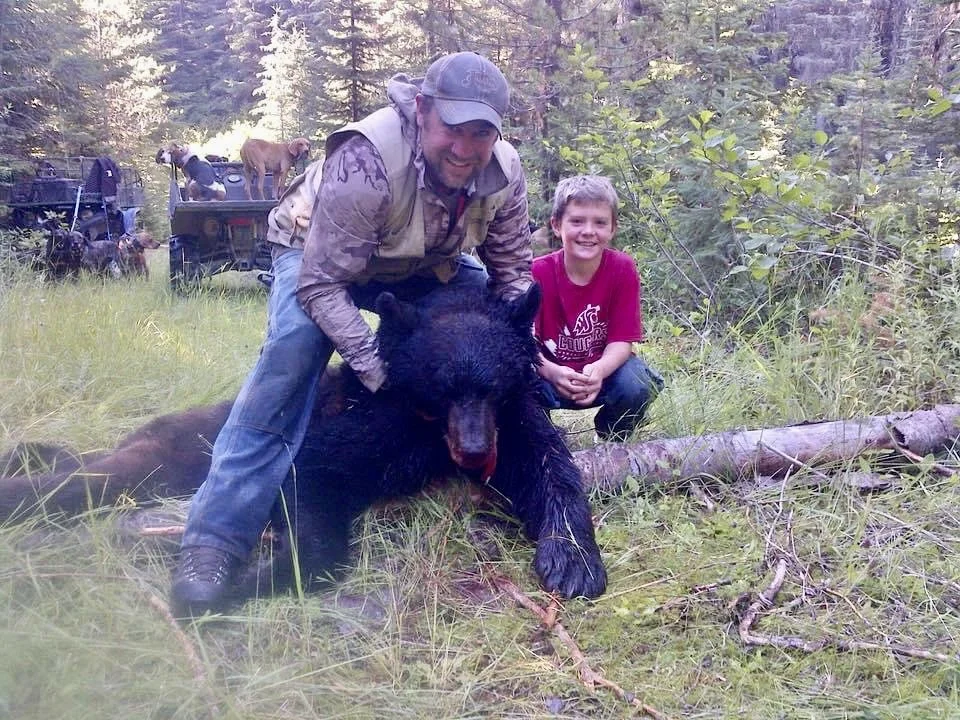 Two men and a young boy kneeling with a large black bear in a wooded forest area.