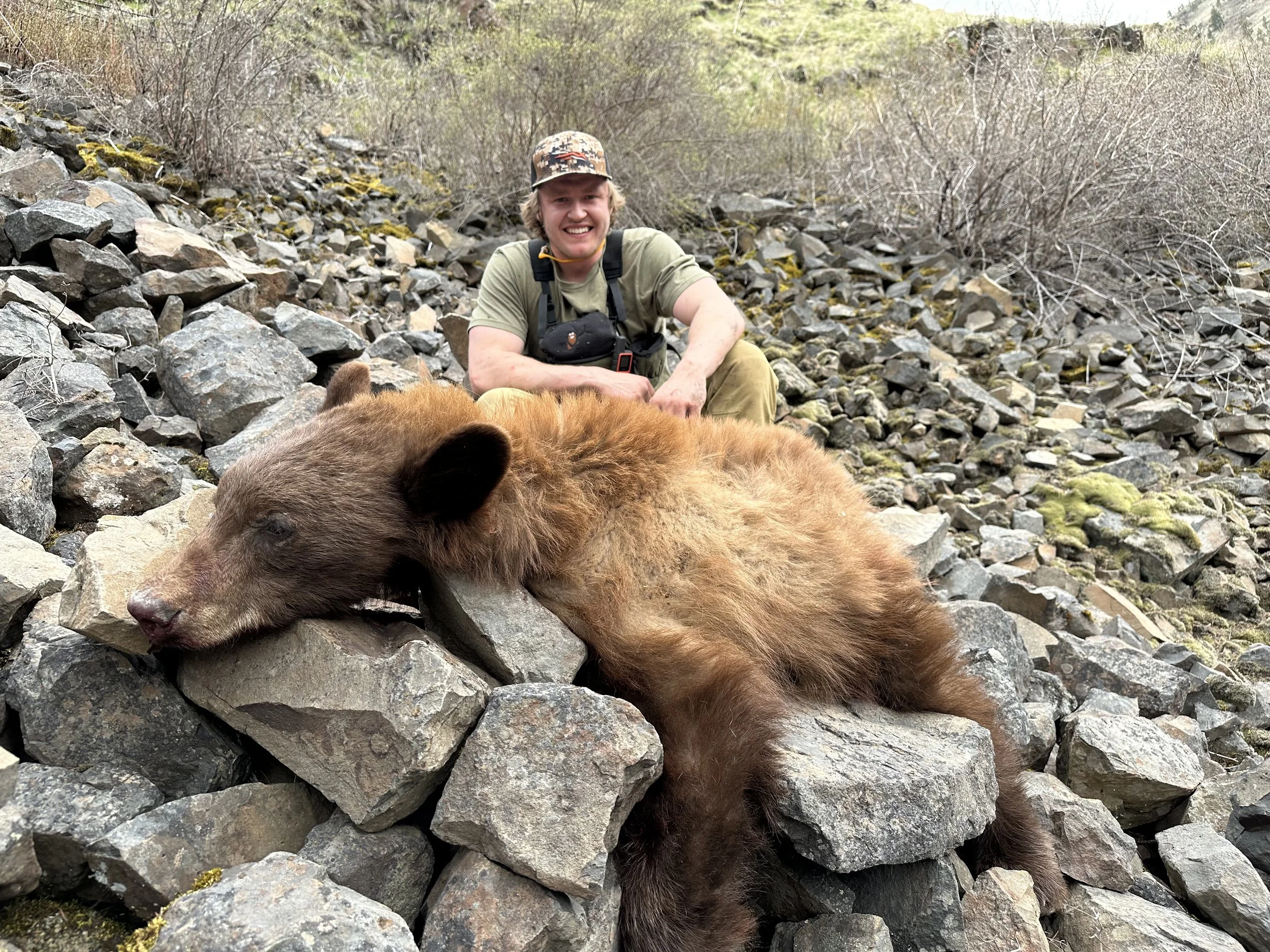 A smiling man in outdoor clothing and a camouflage hat kneels next to a large, bear lying on rocks.