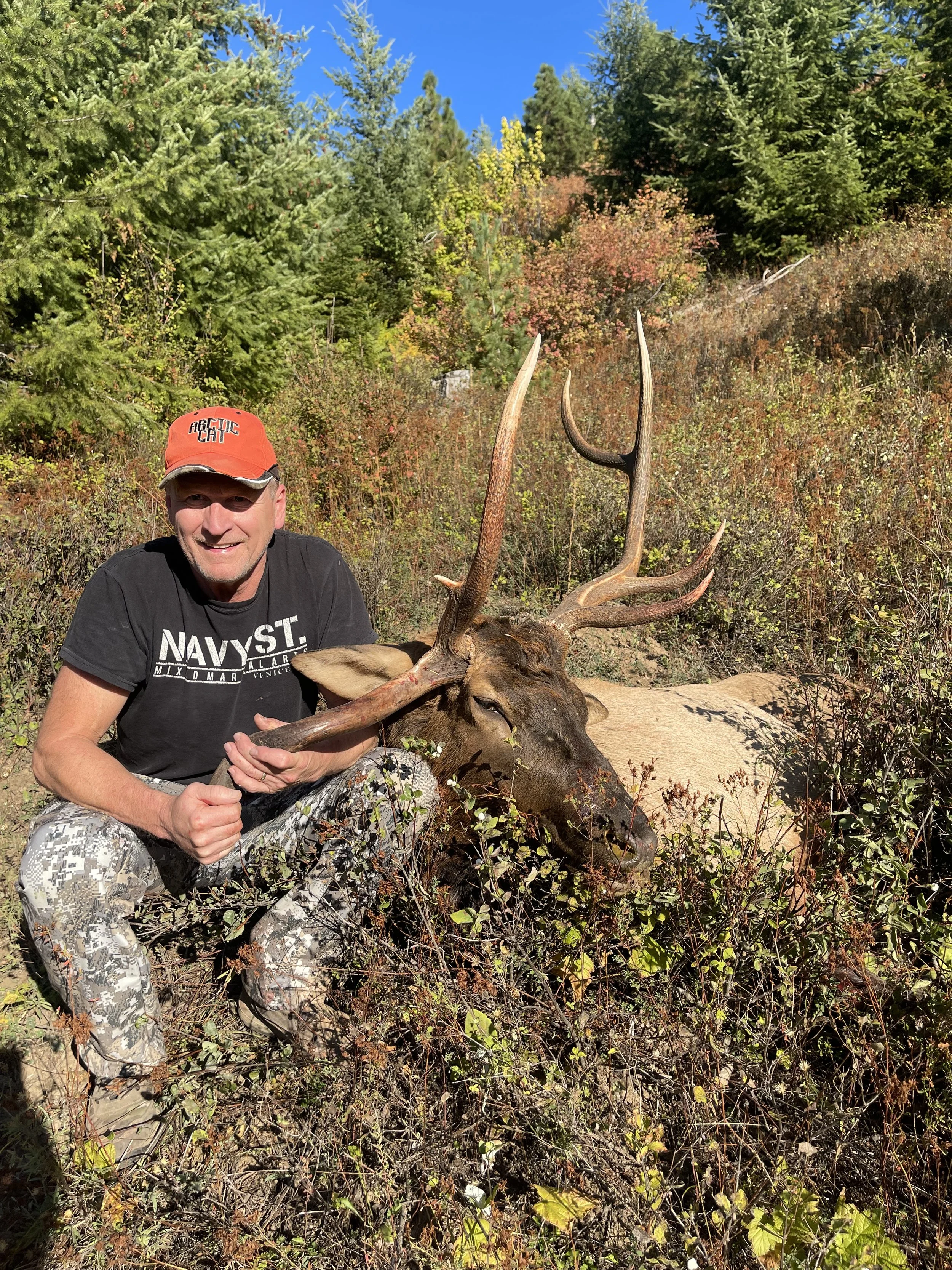 A man in camouflage pants and a black t-shirt holds the antlers of a large elk he has hunted, with a background of trees and bushes.