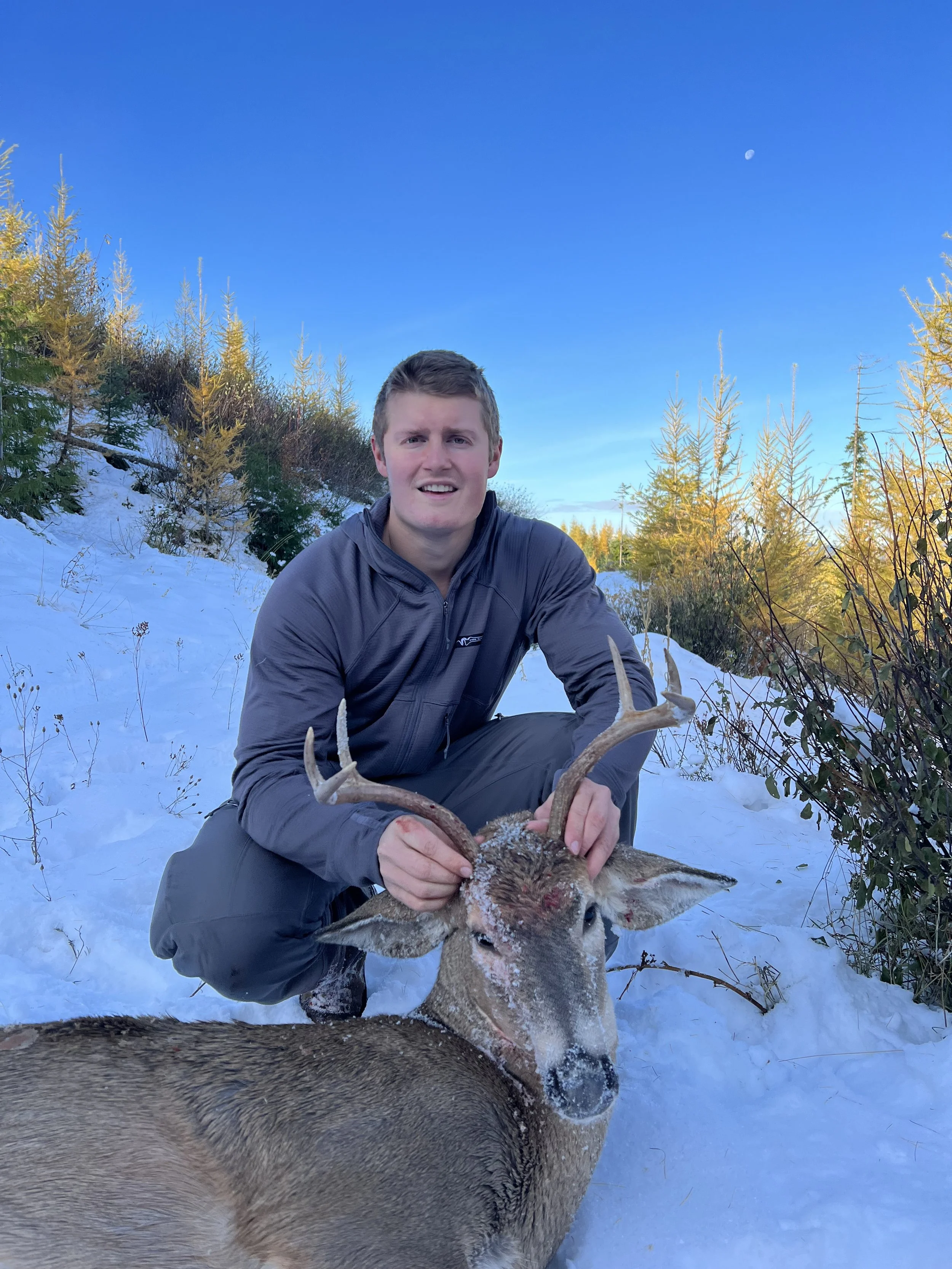 A young man kneeling in the snow, holding the antlers of a recently hunted deer in a snowy wooded area during daytime.