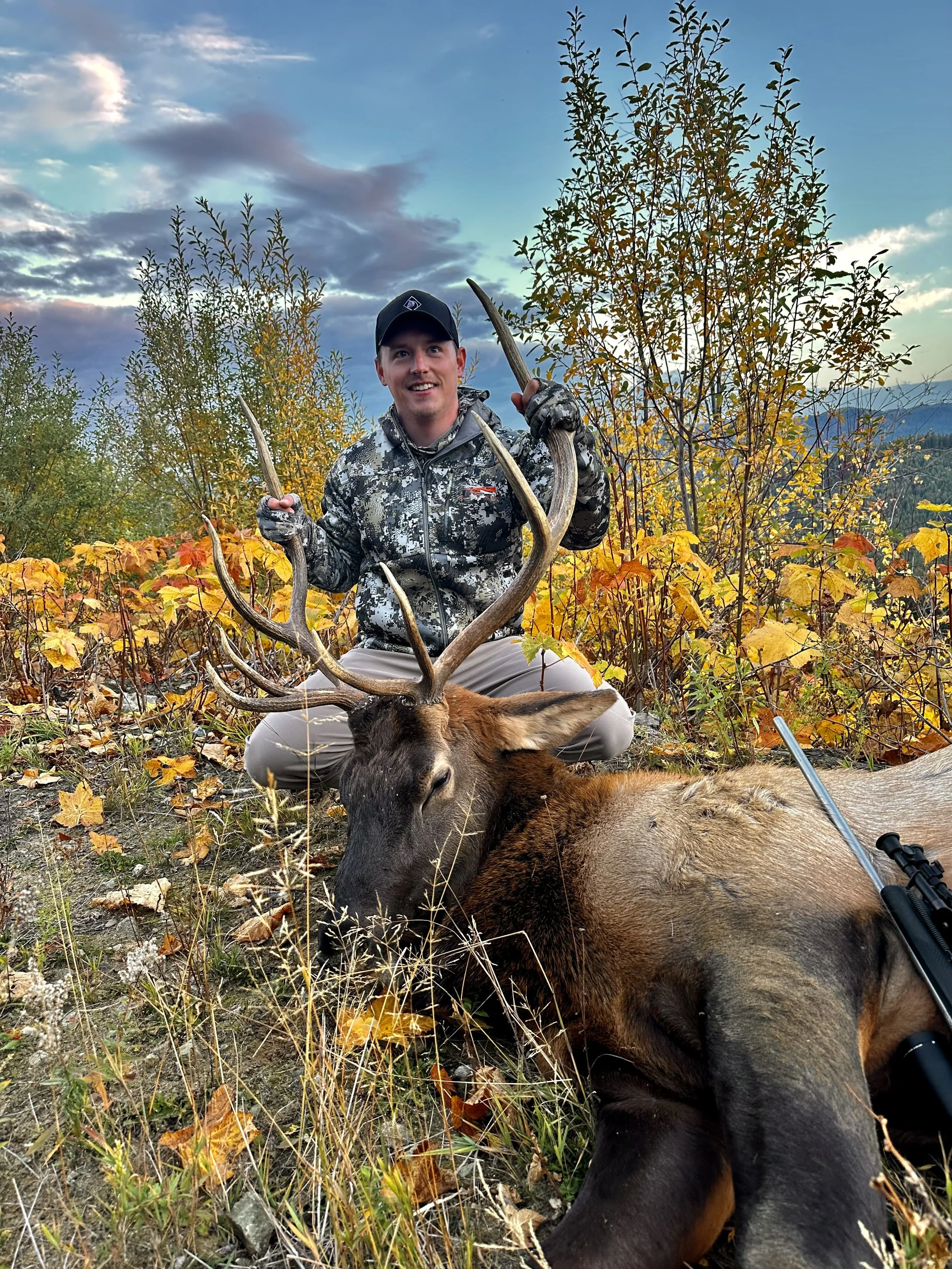 A man in camouflage clothing holding antlers of a large dead elk lying on the ground, outdoors with trees and a cloudy sky in the background.