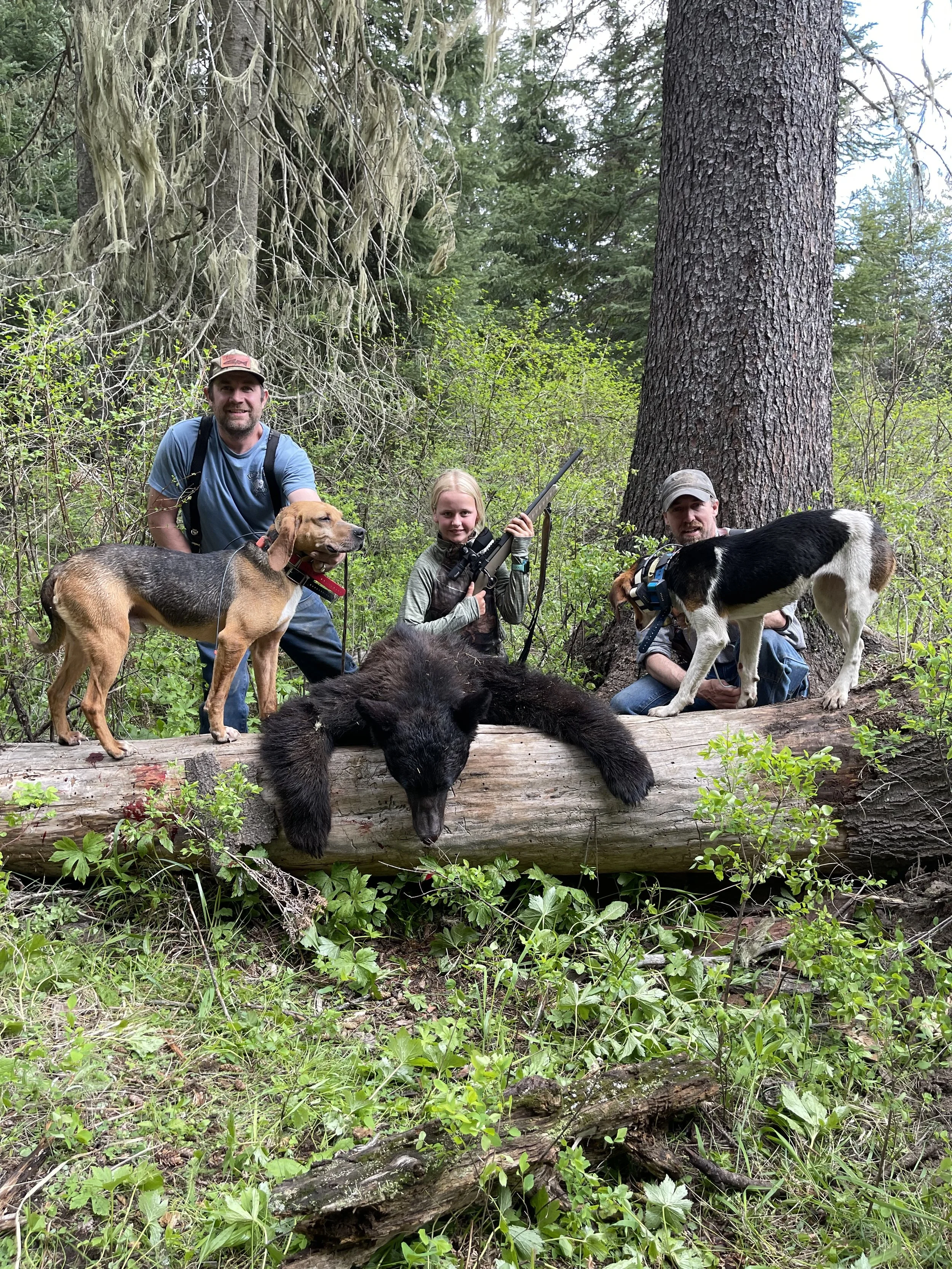 Three people with two dogs and a bear cub in a forest. The man on the right is holding a black and white dog, the girl in the middle is holding a rifle, and the man on the left is standing next to a beagle dog. All three are outdoors among trees and 