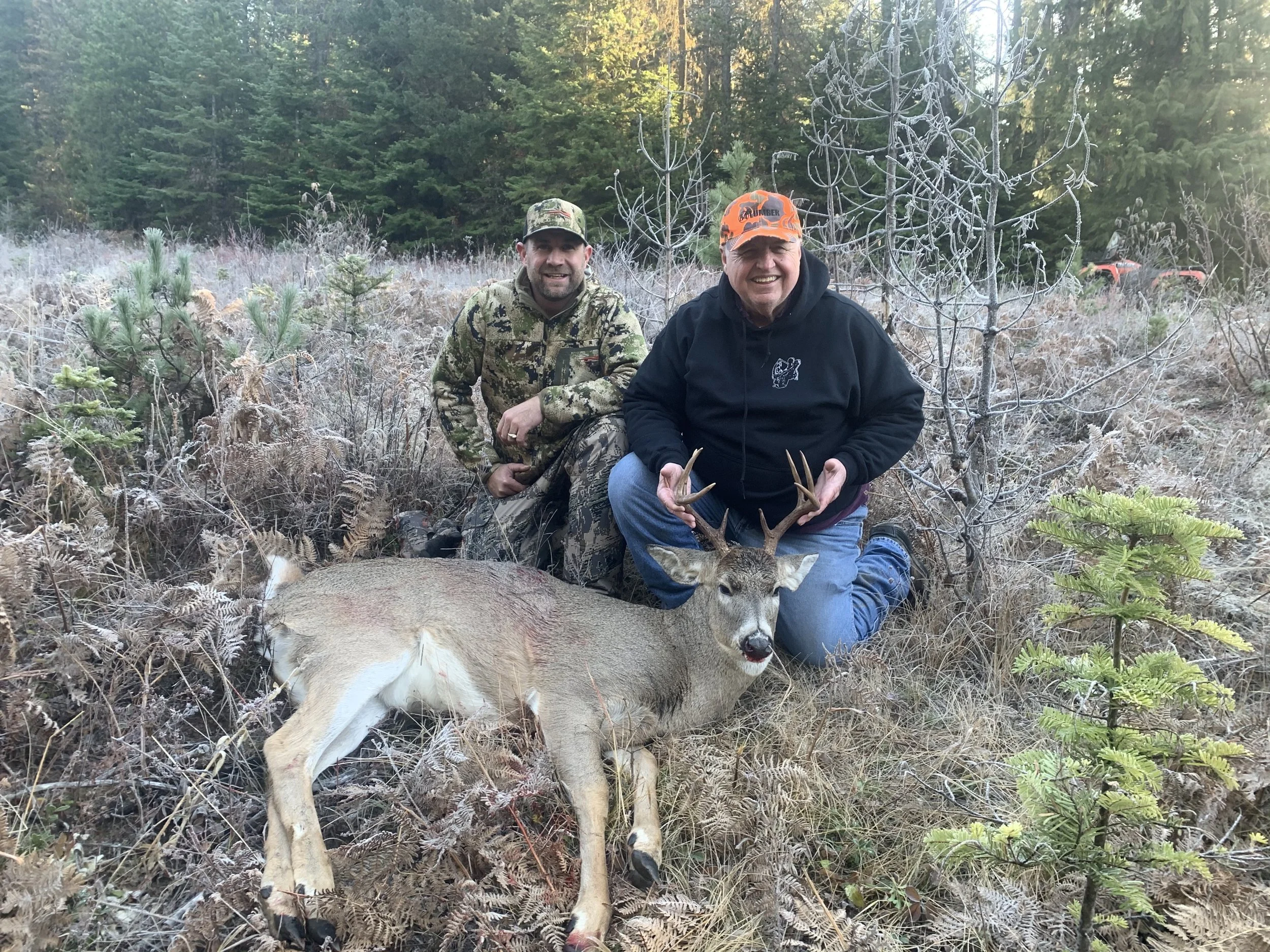 Two men kneeling in a forested area with a dead deer, holding the deer's antlers and smiling at the camera.