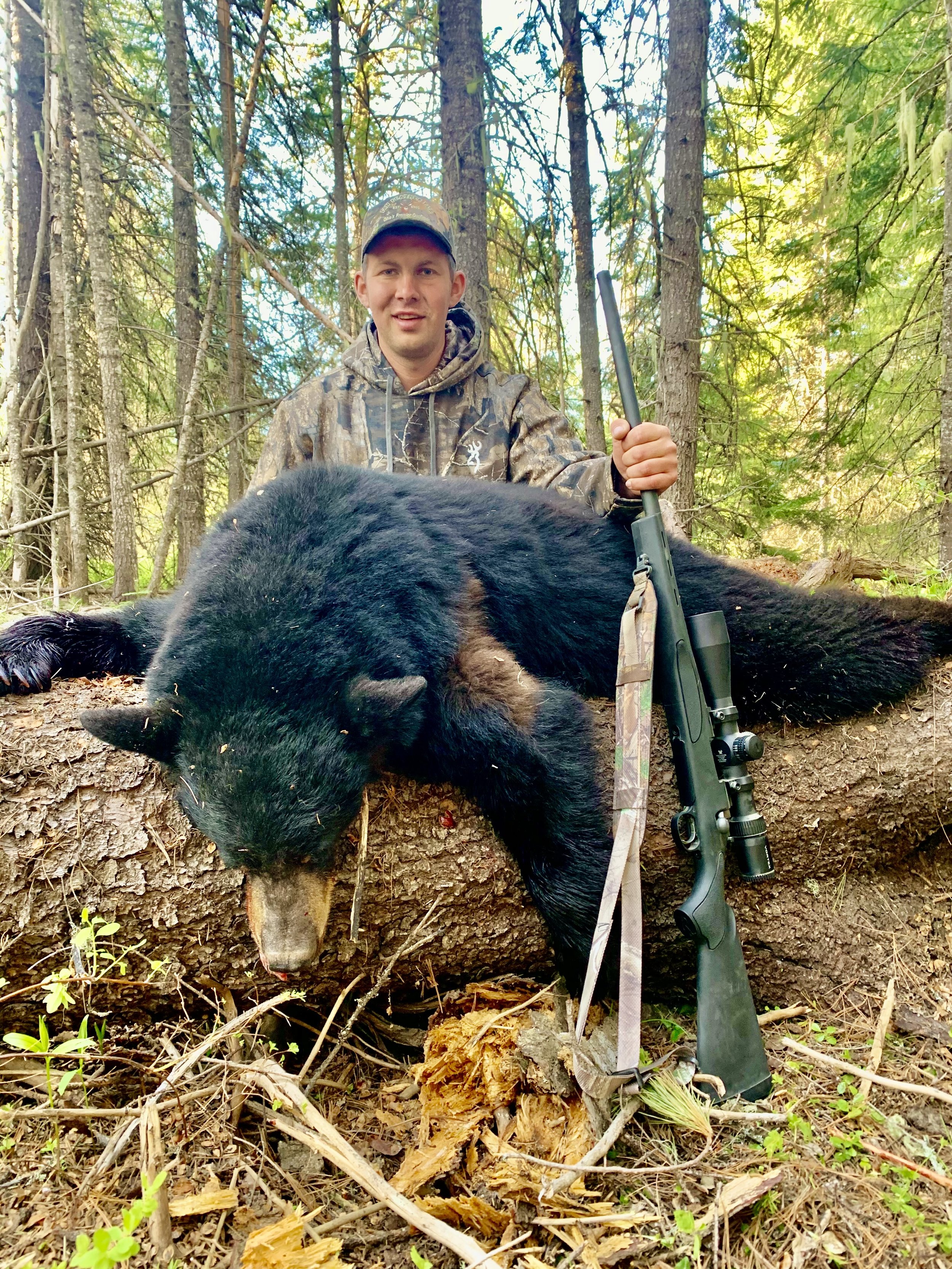 A man dressed in camouflage outdoors beside a large fallen tree trunk, holding a rifle, with a large black bear laying on the tree trunk.