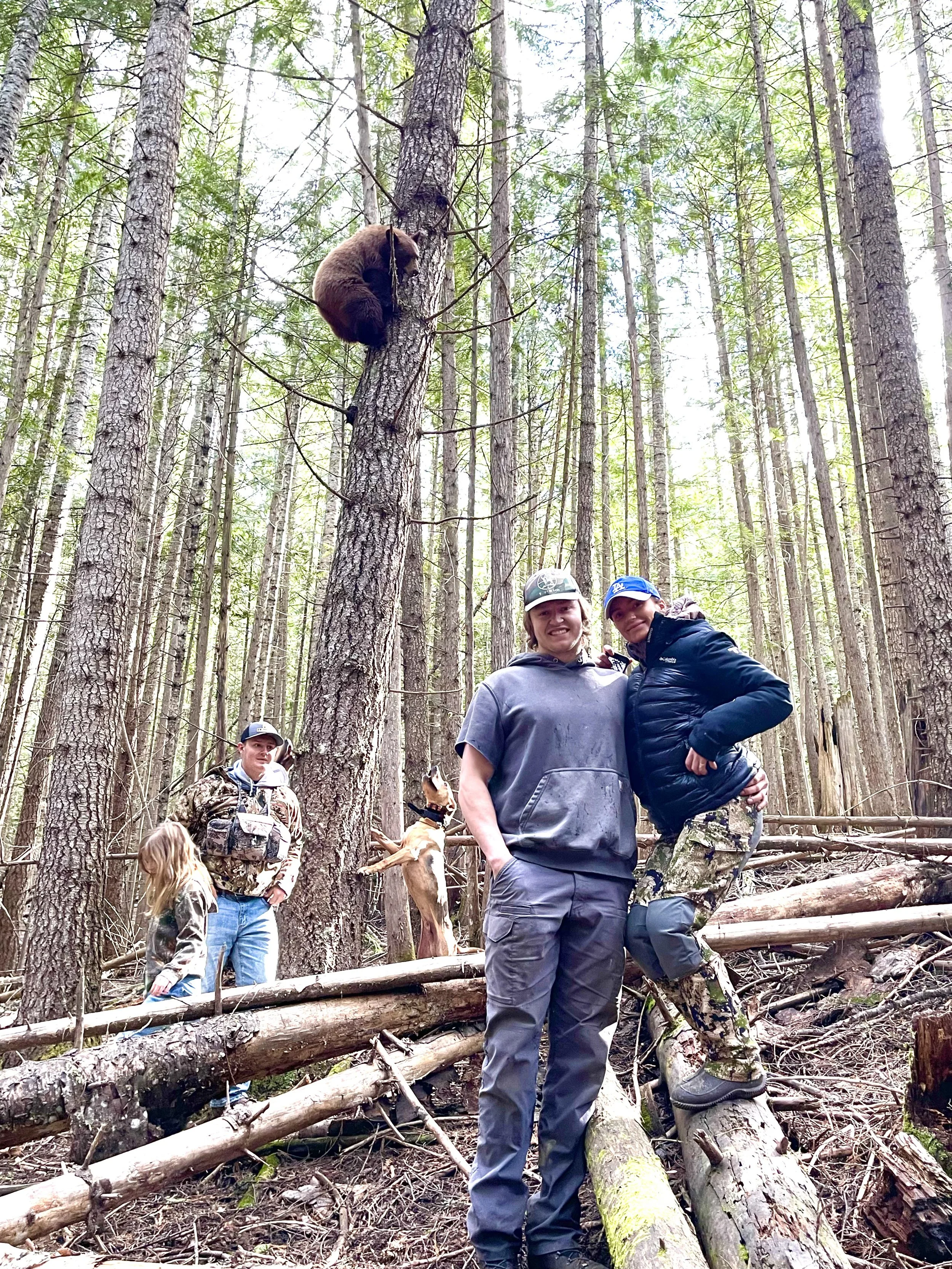 Group of people in a forest with tall trees, two women in the foreground smiling and posing, one wearing camouflage pants, others with casual outdoor clothing, a dog on a leash, and a bear climbing a tree in the background.