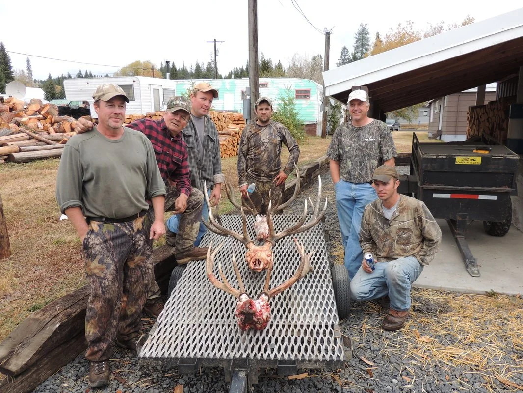 Six men standing outdoors behind a deer with large antlers that has been hunted, with carved head and antlers placed on a metal grate on a trailer. The men are dressed in camouflage and outdoor clothing, and the background shows trees, a trailer, and