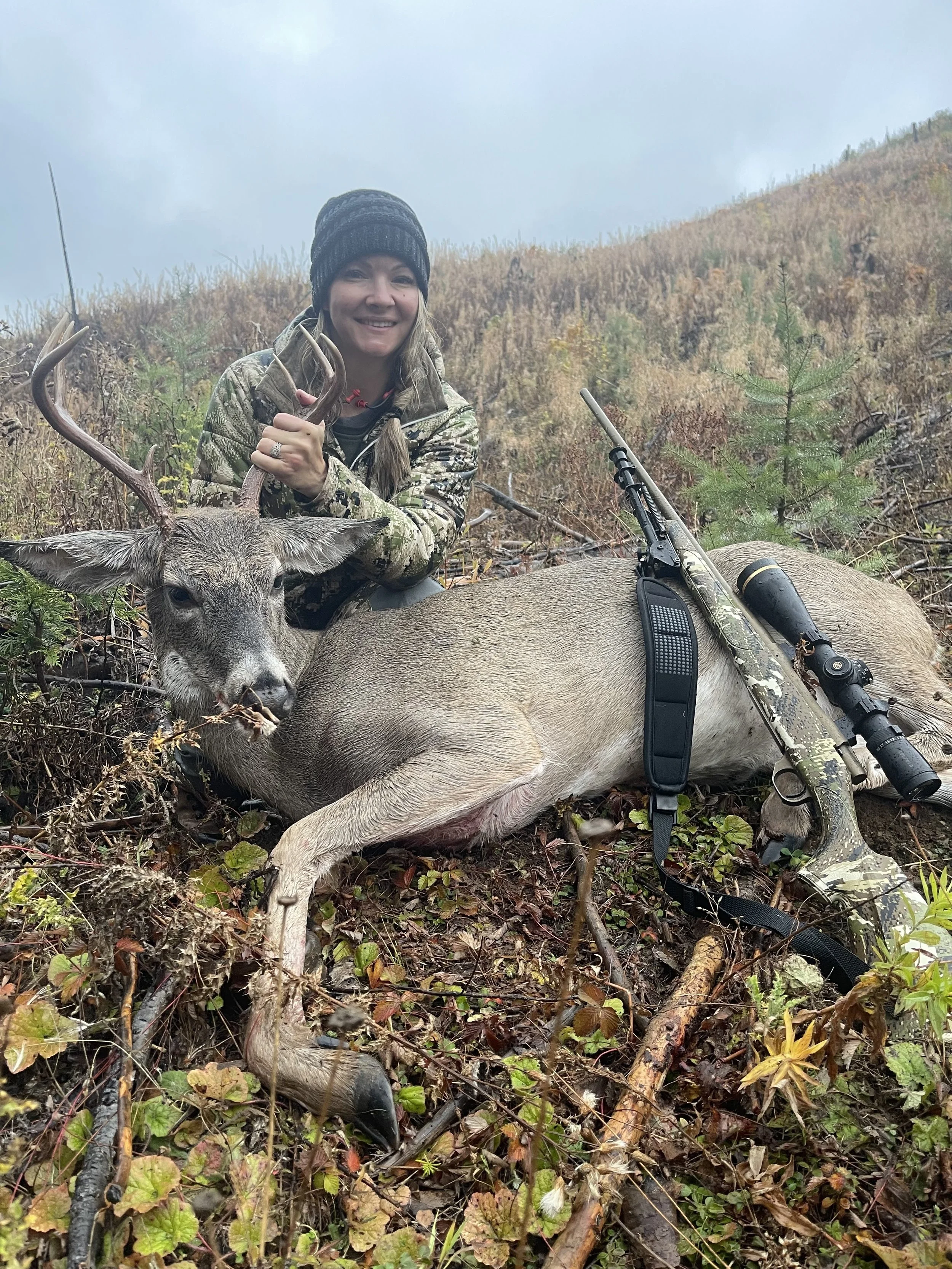 A woman in camouflage clothing and a black beanie smiling and kneeling next to a large, freshly hunted deer with antlers. The deer is lying on the ground among foliage, and a scoped rifle is resting on the deer's body in a forested outdoor setting.