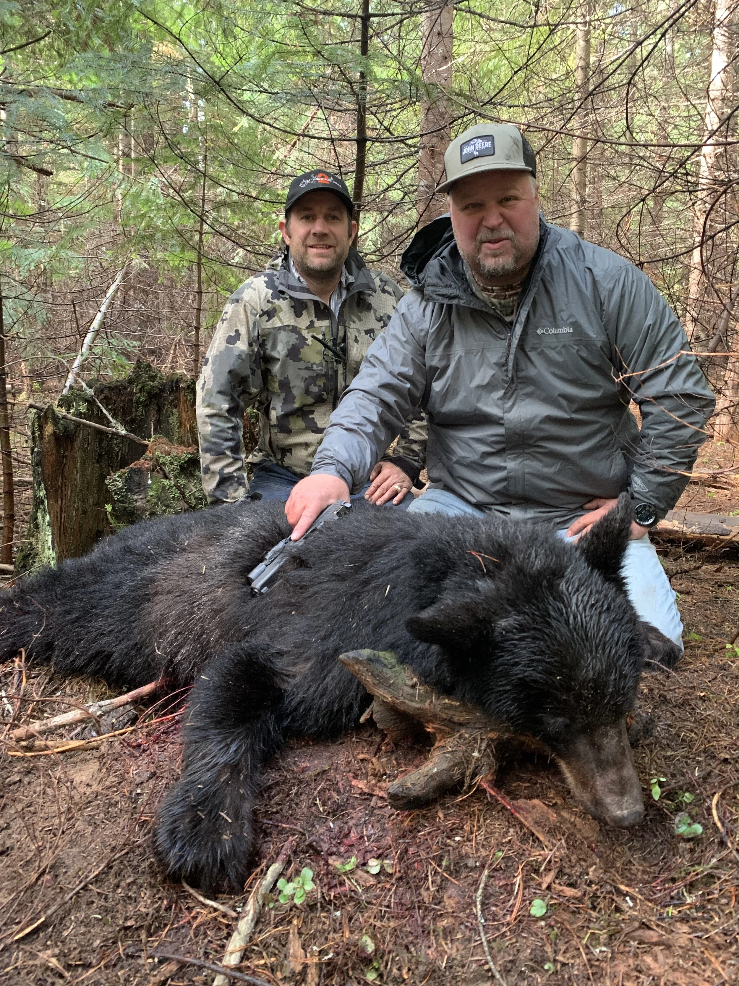 Two men kneeling in a forest, posing with a large deceased black bear lying on the ground. One man has a handgun resting on the bear, and they both appear proud of their hunt.