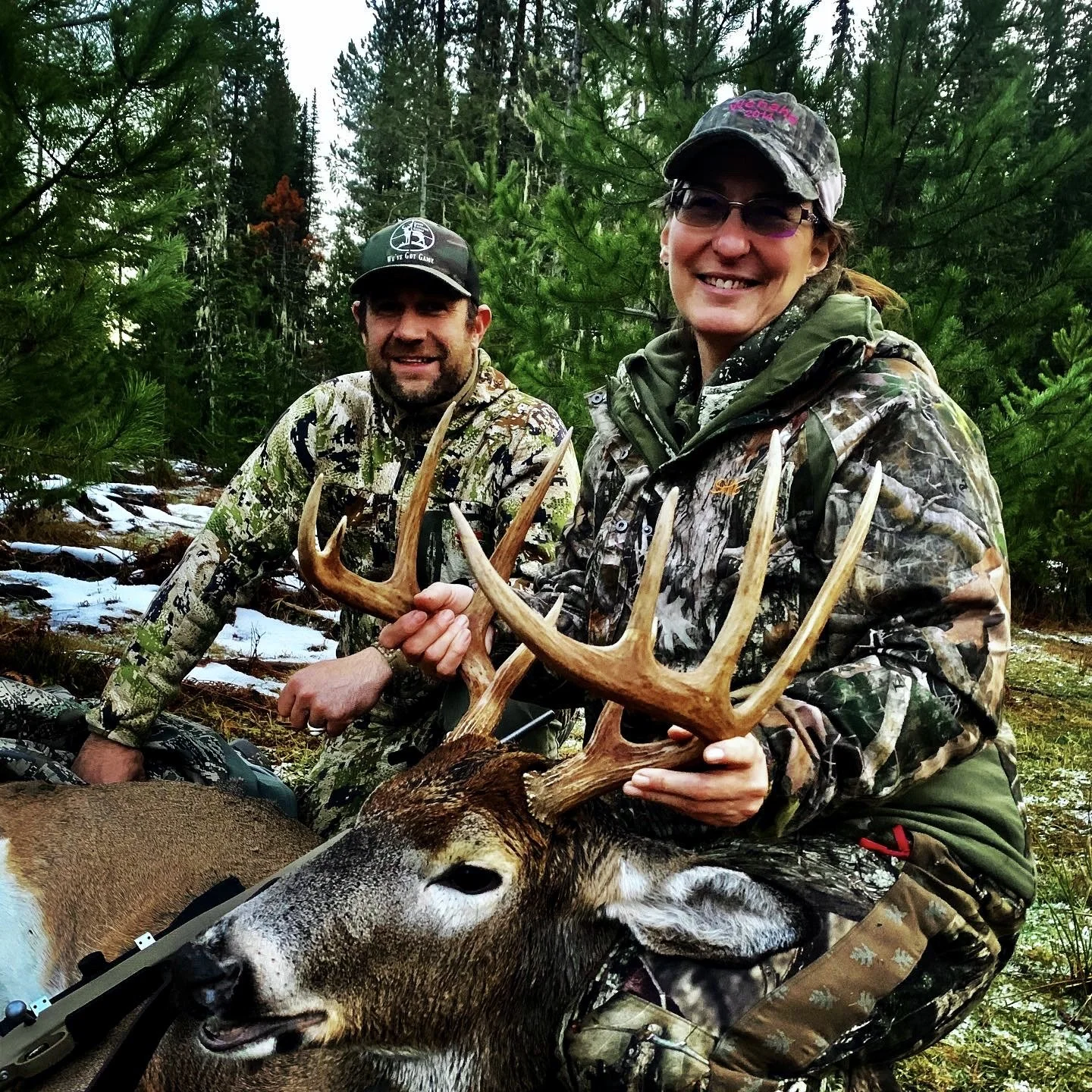 A woman and man in camouflage hunting gear outdoors in a forest, holding a large deer with antlers, smiling at the camera.