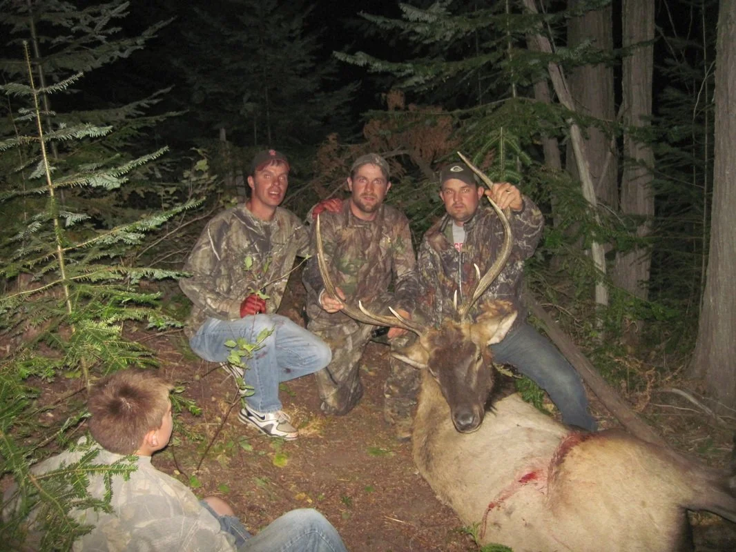 Three hunters pose with a large elk they have hunted, holding its antlers in a forest at night, with two young boys sitting nearby.