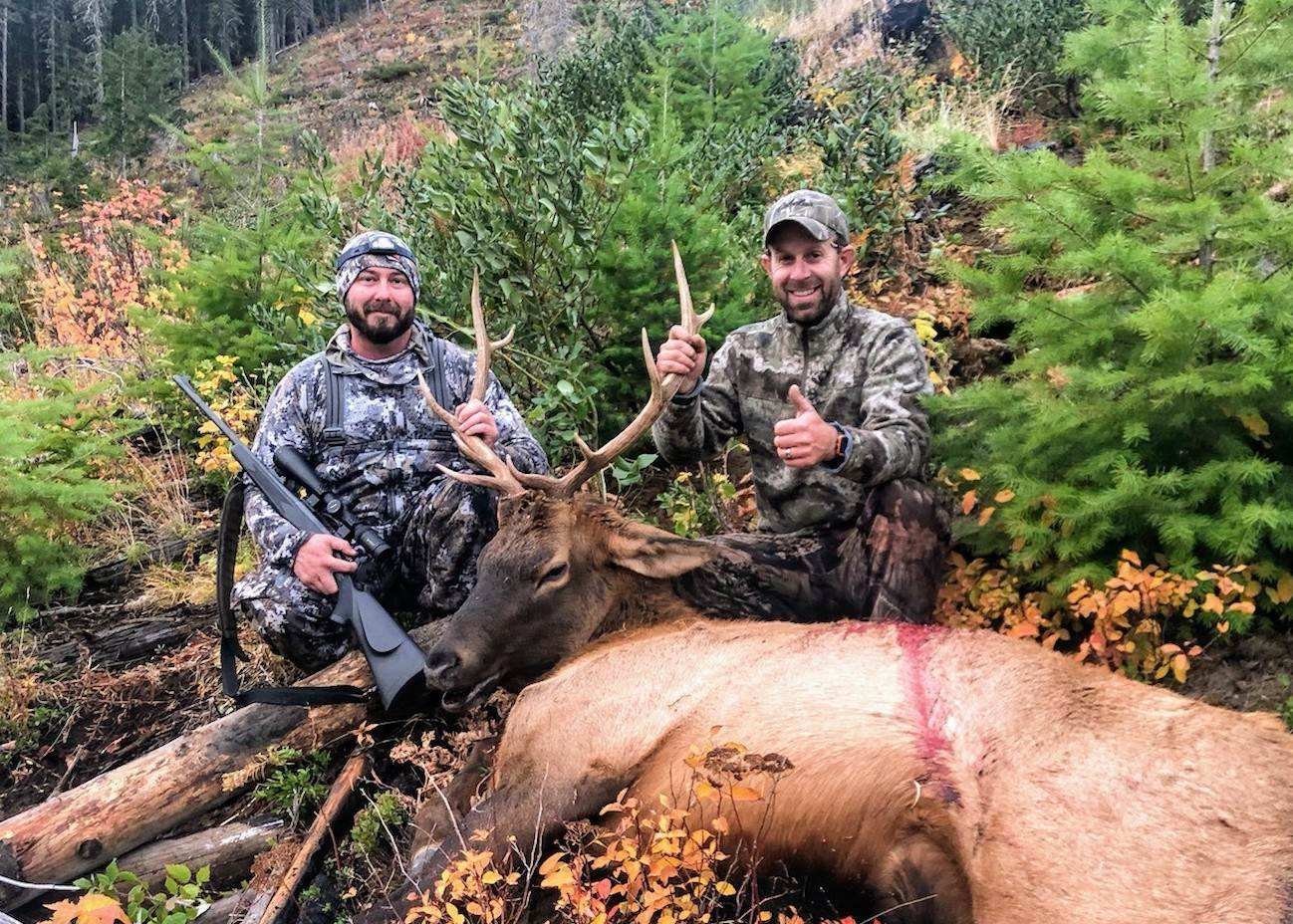 Two men in camouflage clothing kneel behind a large elk with impressive antlers, which lies on the ground with a visible wound. One man is holding a rifle, and the other shows a thumbs-up. They are in a forested area with green trees and shrubs.