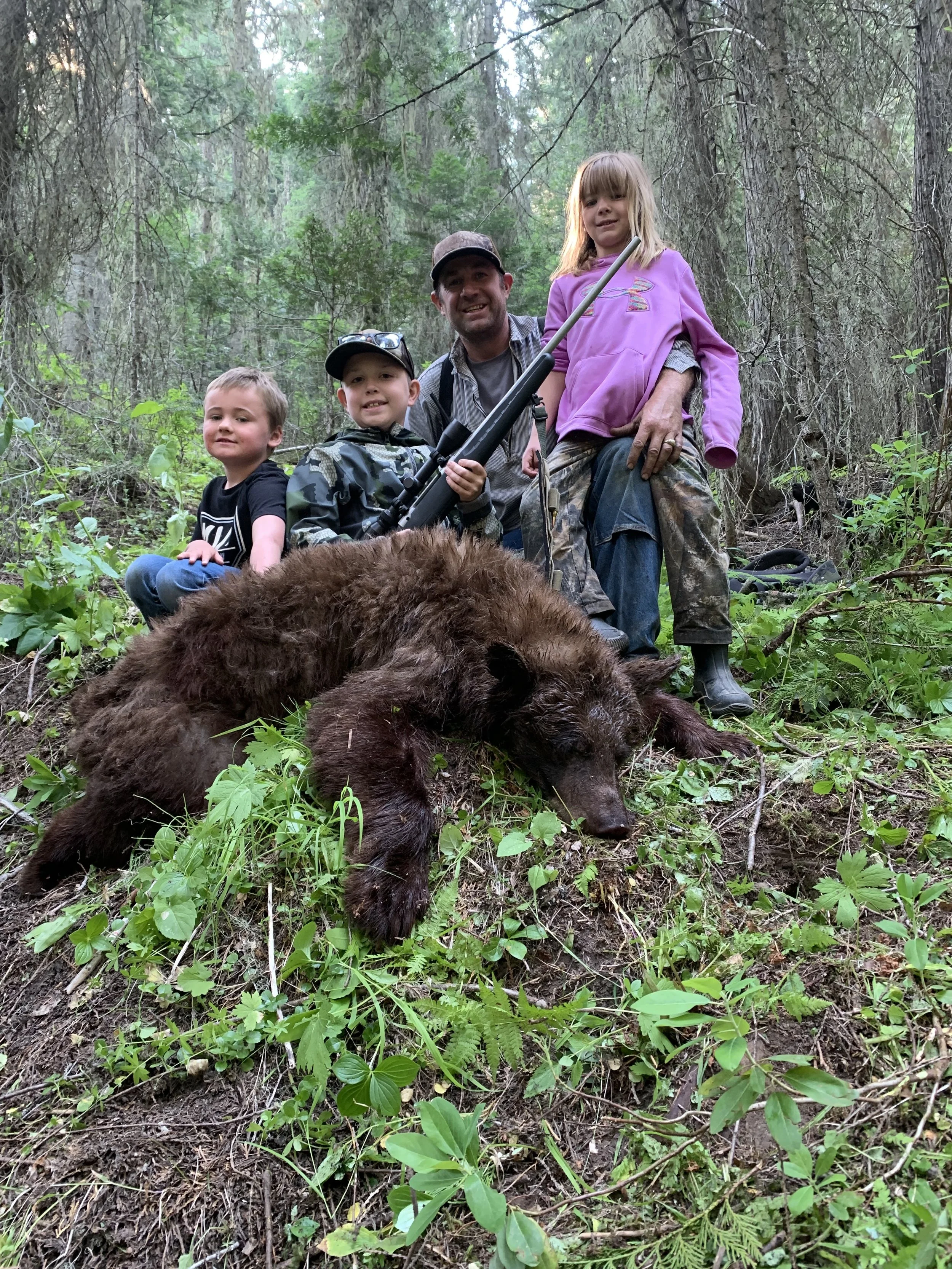 A man and three children in a forest, posing with a large dead bear on the ground. The man is holding a rifle, and the children are smiling, with one child sitting on the ground and another standing next to the man. The scene is set in a densely wood