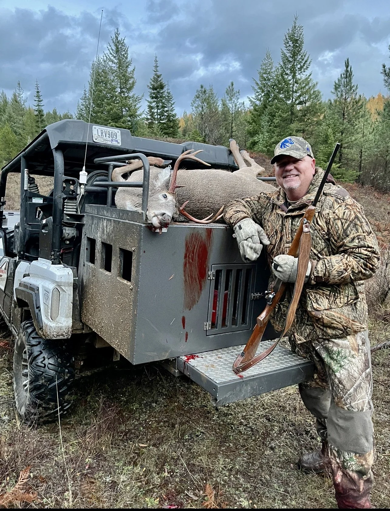 A man in camouflage clothing and a cap, smiling, stands next to a hunting vehicle with a rifle. The vehicle carries a deer with antlers and a sheep or goat. The scene is outdoors with trees and cloudy sky.