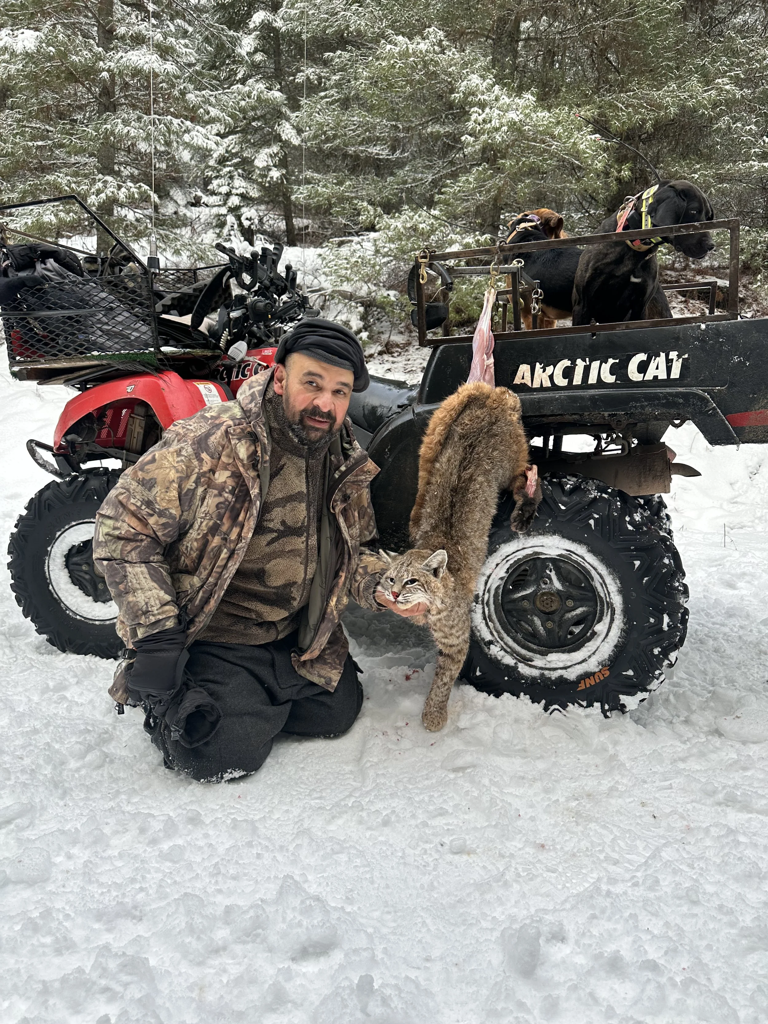 A man in camouflage clothing kneeling in snow, holding a dead animal, with a lynx in front and a black dog in the background, near a black ATV labeled 'Arctic Cat' in a snowy forest.