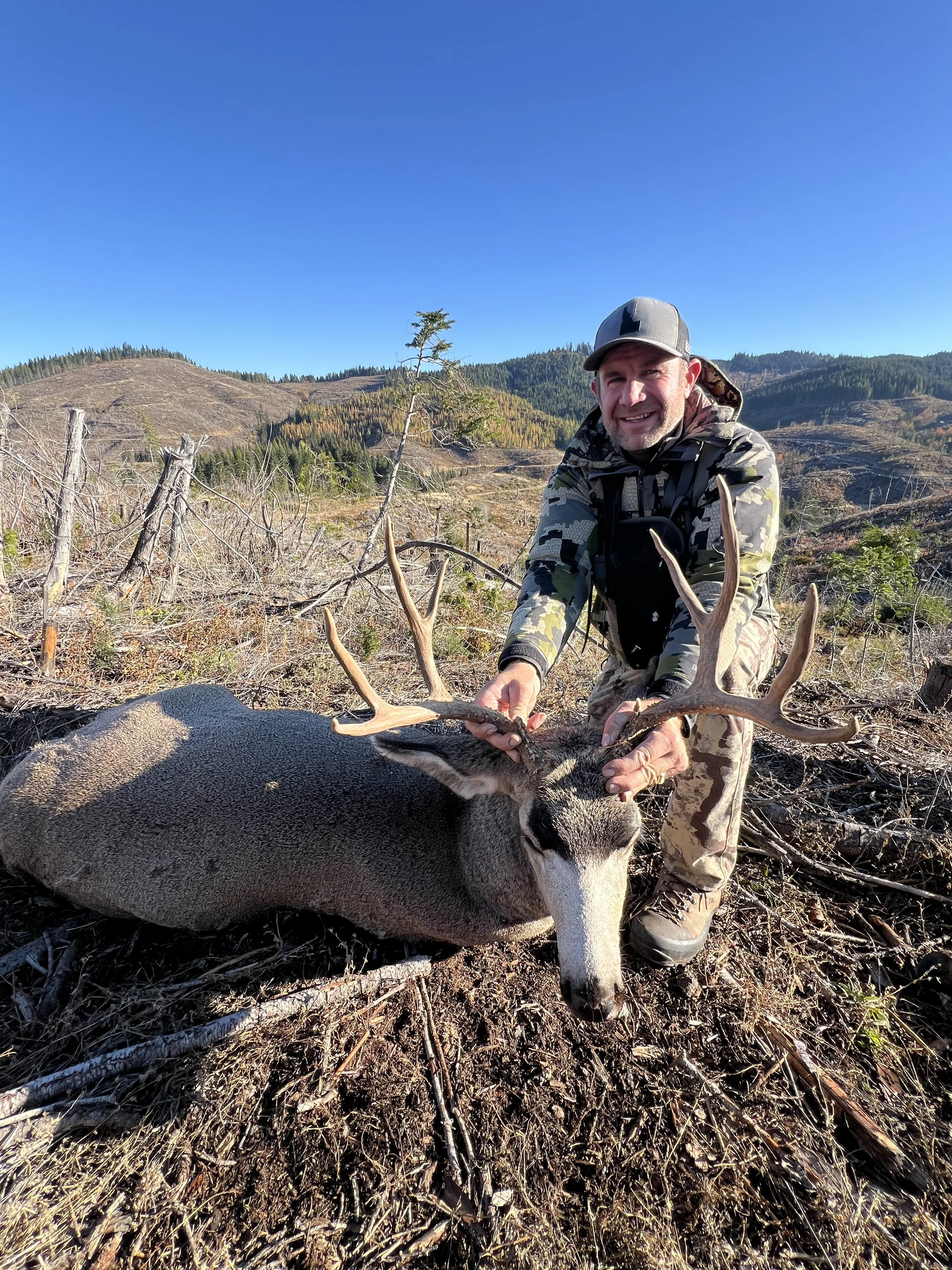 A man in camouflage clothing and a gray hat kneels beside a large mule deer with antlers, smiling at the camera in a mountainous, forested landscape.