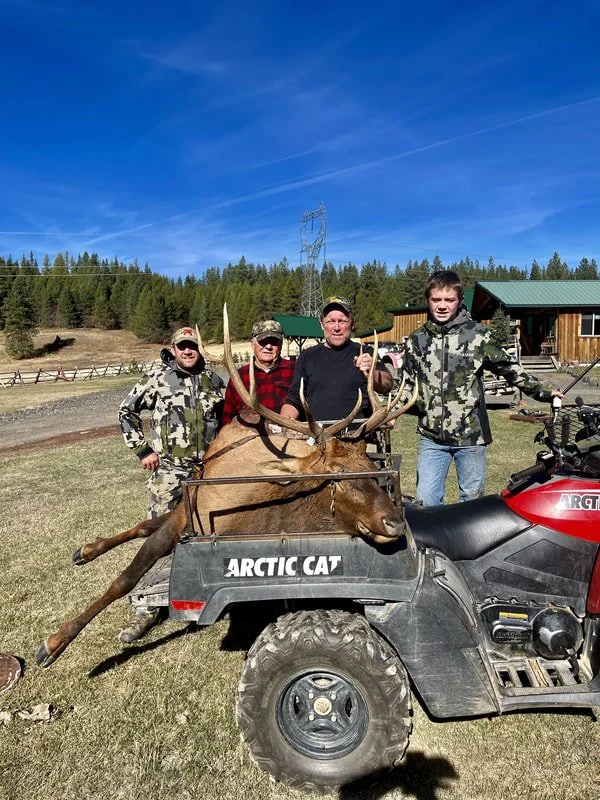 Four men standing together outdoors with a large elk in a cart labeled 'Arctic Cat.' The elk is lying down with its antlers displayed, and the men appear proud. The background features a green forest, blue sky, and some buildings.