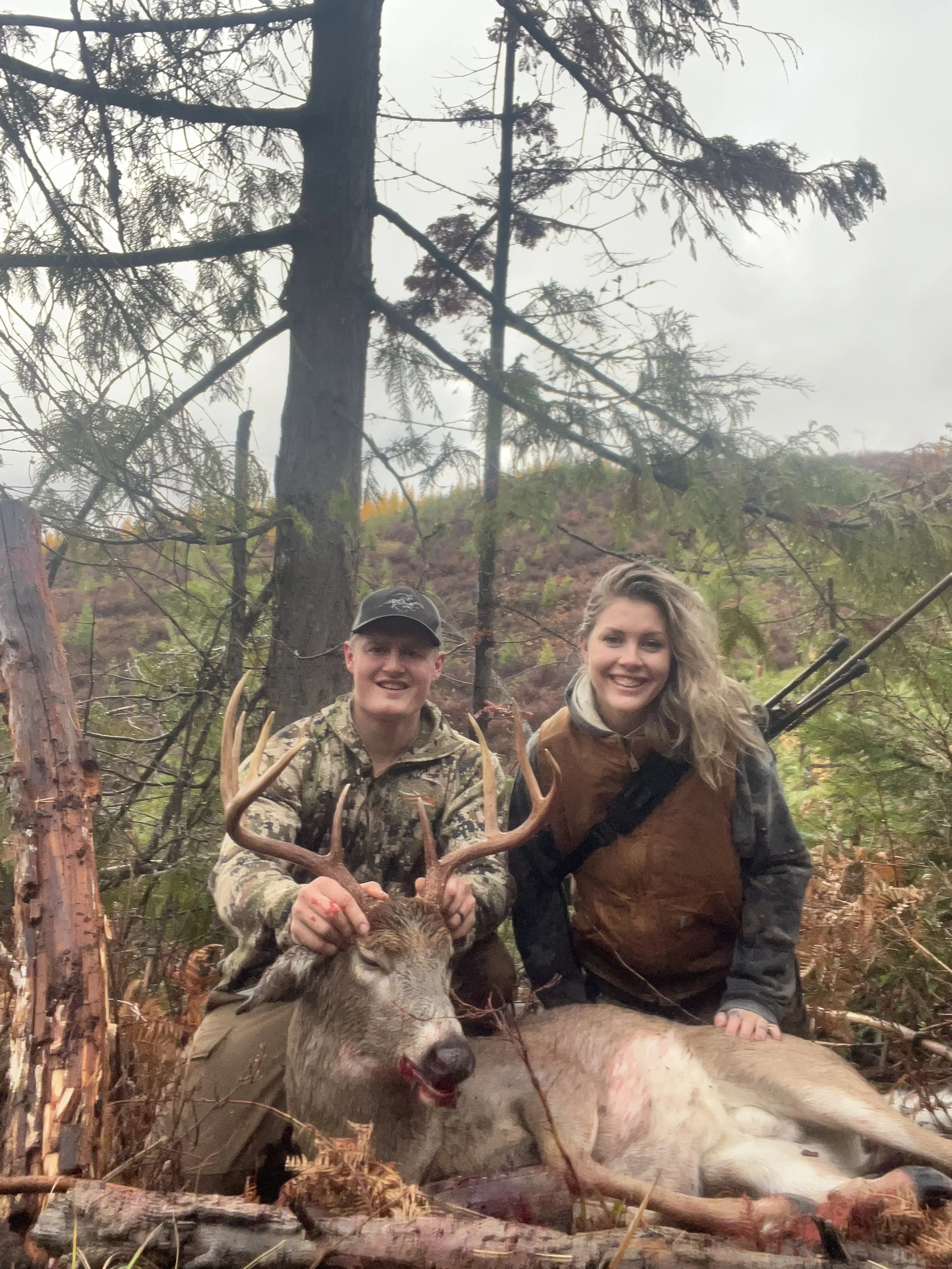 Two smiling hunters posing with a large deer they have hunted, in a forested area during daytime.