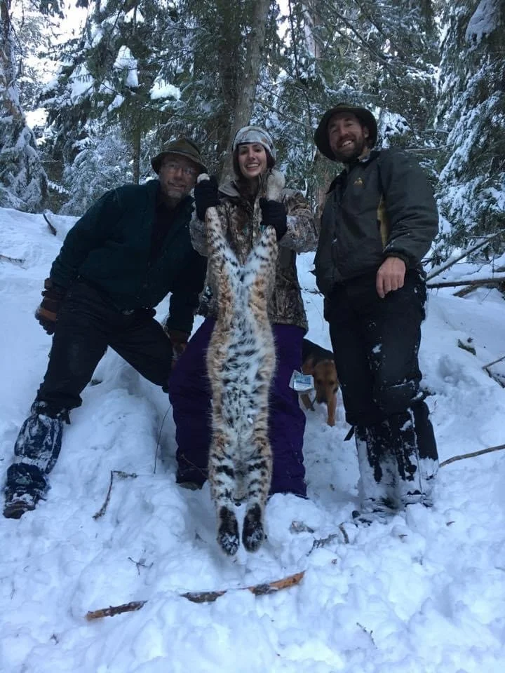 Three people in winter clothing standing in snow-covered forest, holding a large snow leopard pelt.