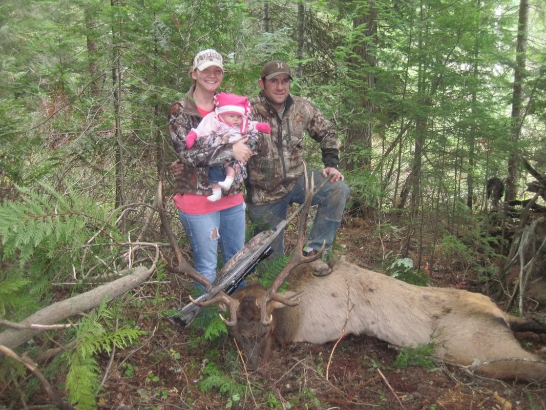A family of three, a man, a woman, and a baby, posing in a forest with a large, elk with antlers. The woman is holding the baby, and both adults are wearing camouflage clothing. The elk is lying on the ground among trees and greenery.