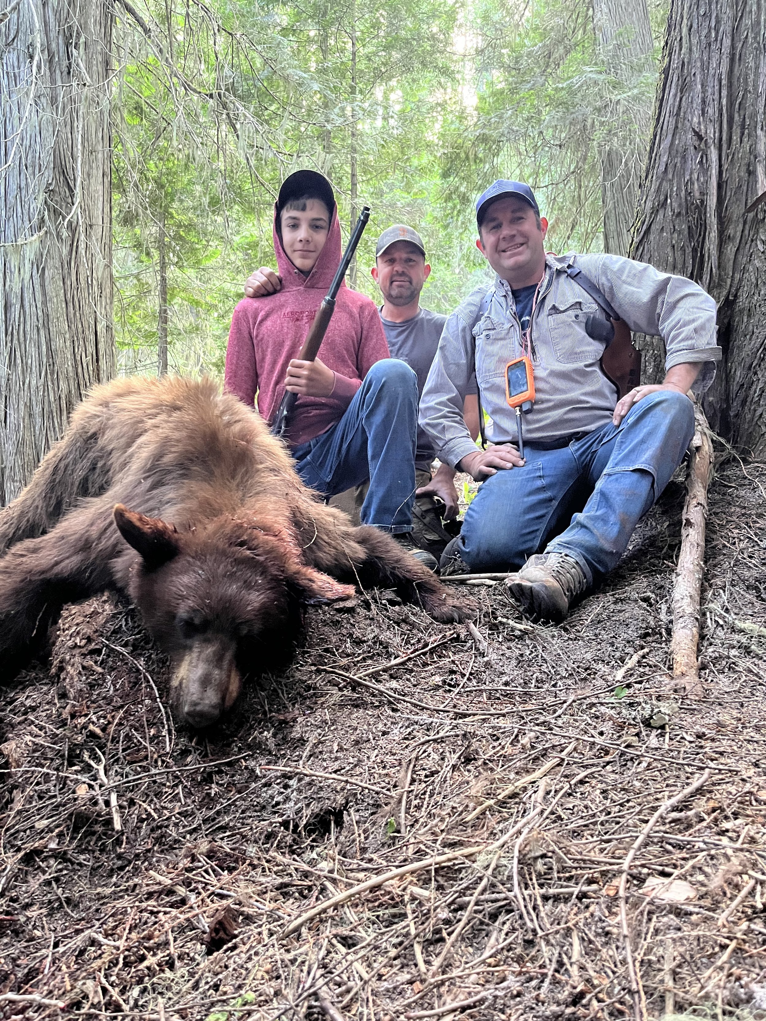 Three people in a forest posing with a dead bear lying on the ground. The group includes a child holding a rifle and two adults, one wearing a cap and the other with a camera or GPS device hanging from his neck.