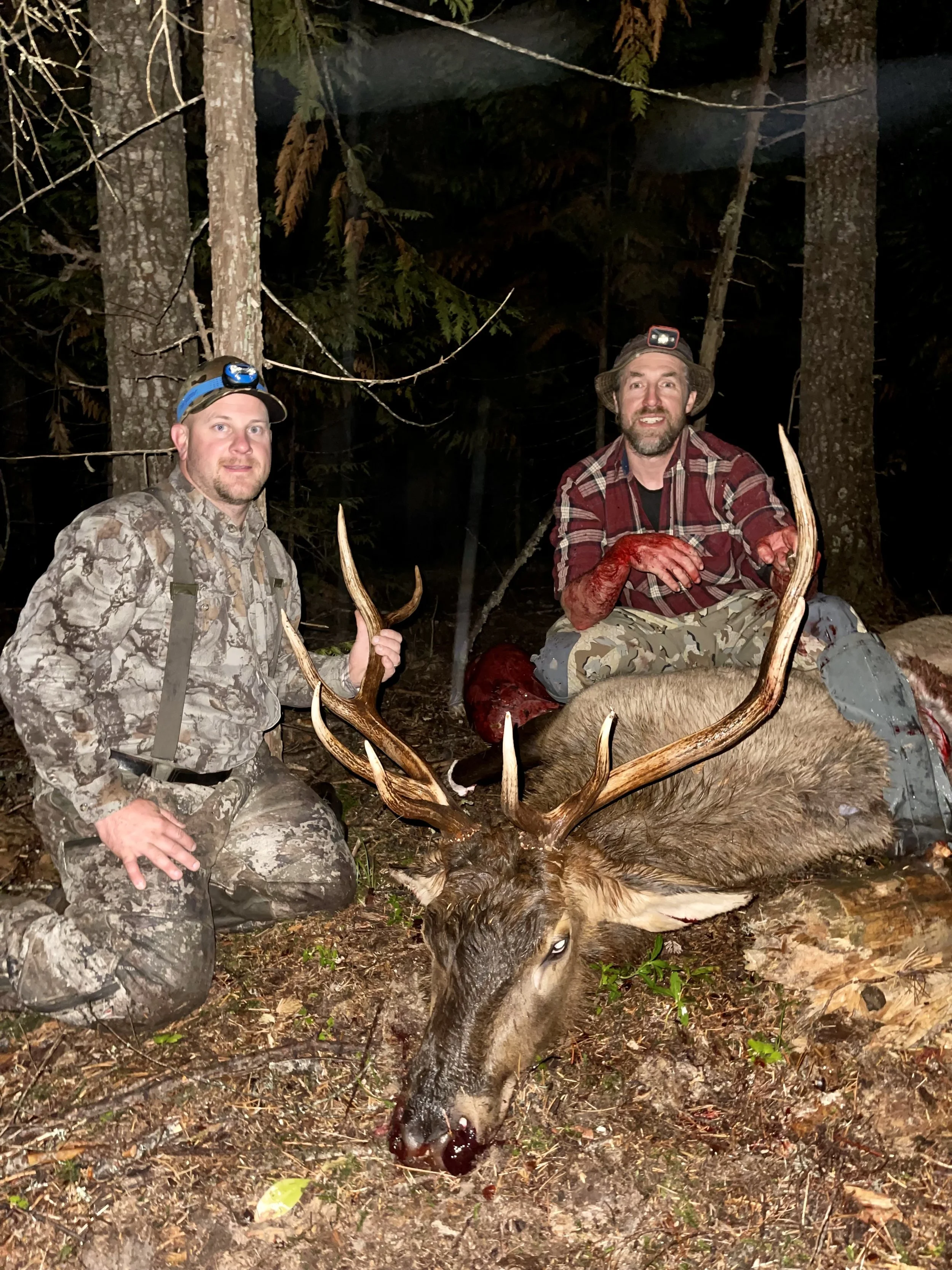 Two men in camouflage and plaid shirts posing with a large deceased deer with big antlers in a forested area at night.