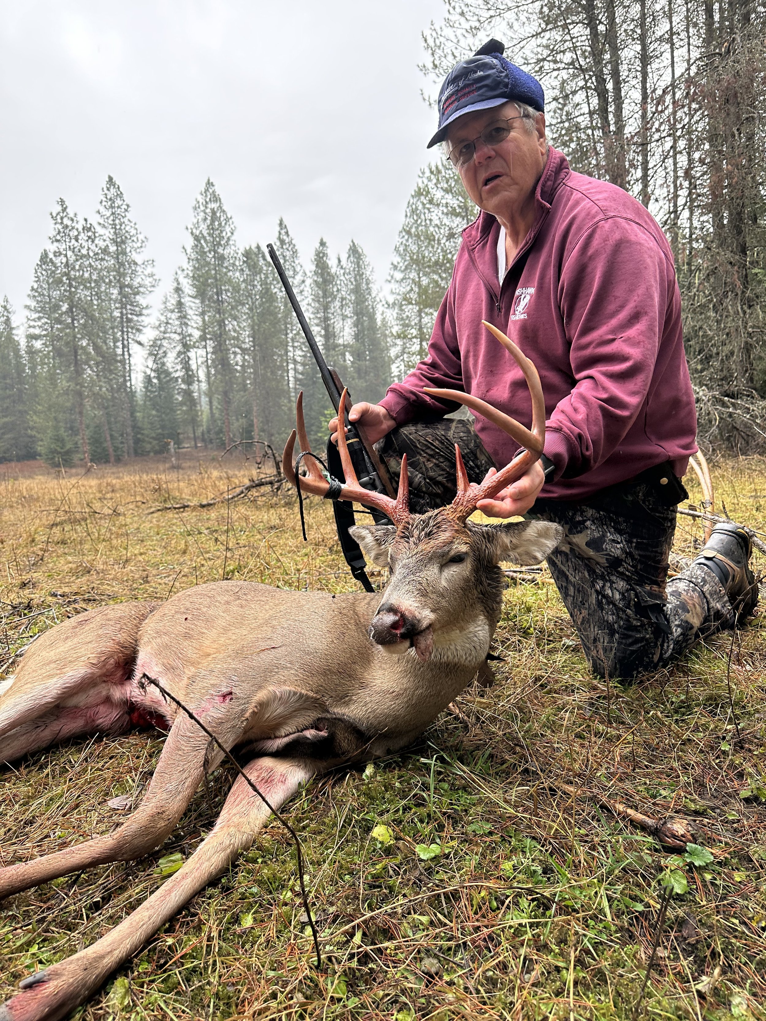 An older man in outdoor clothing kneels on the ground in a forested area, holding a rifle, next to a large deer with antlers lying on the grass, indicating a hunting scene.
