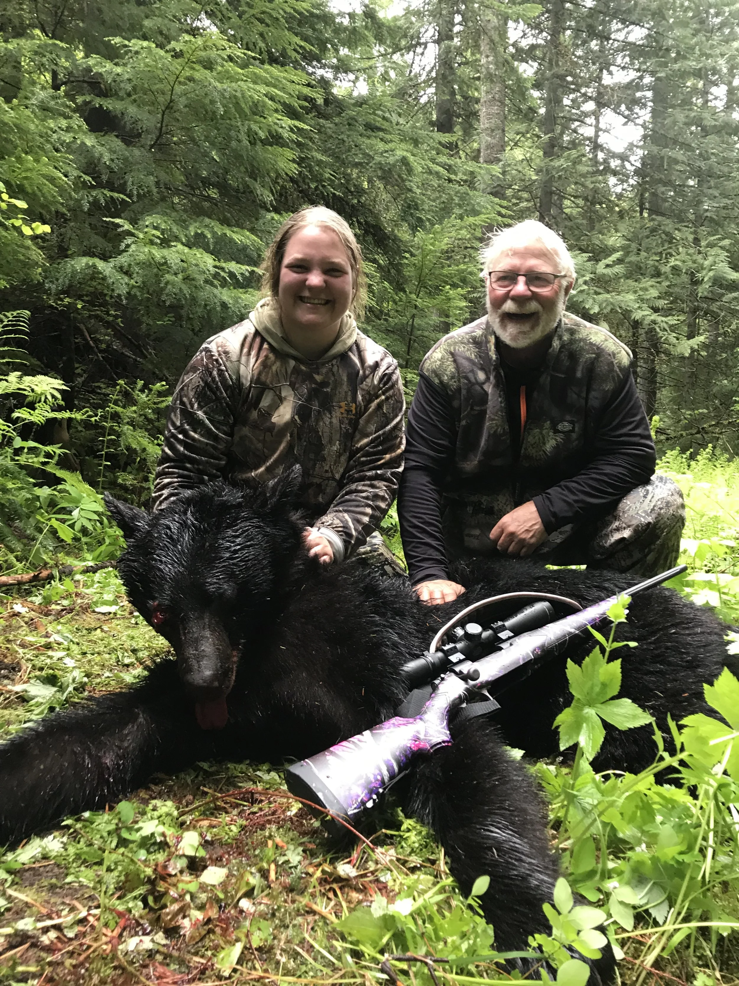 Two people in camouflage clothing kneeling in a forest beside a large, dead black bear with a mounted rifle resting on the bear.