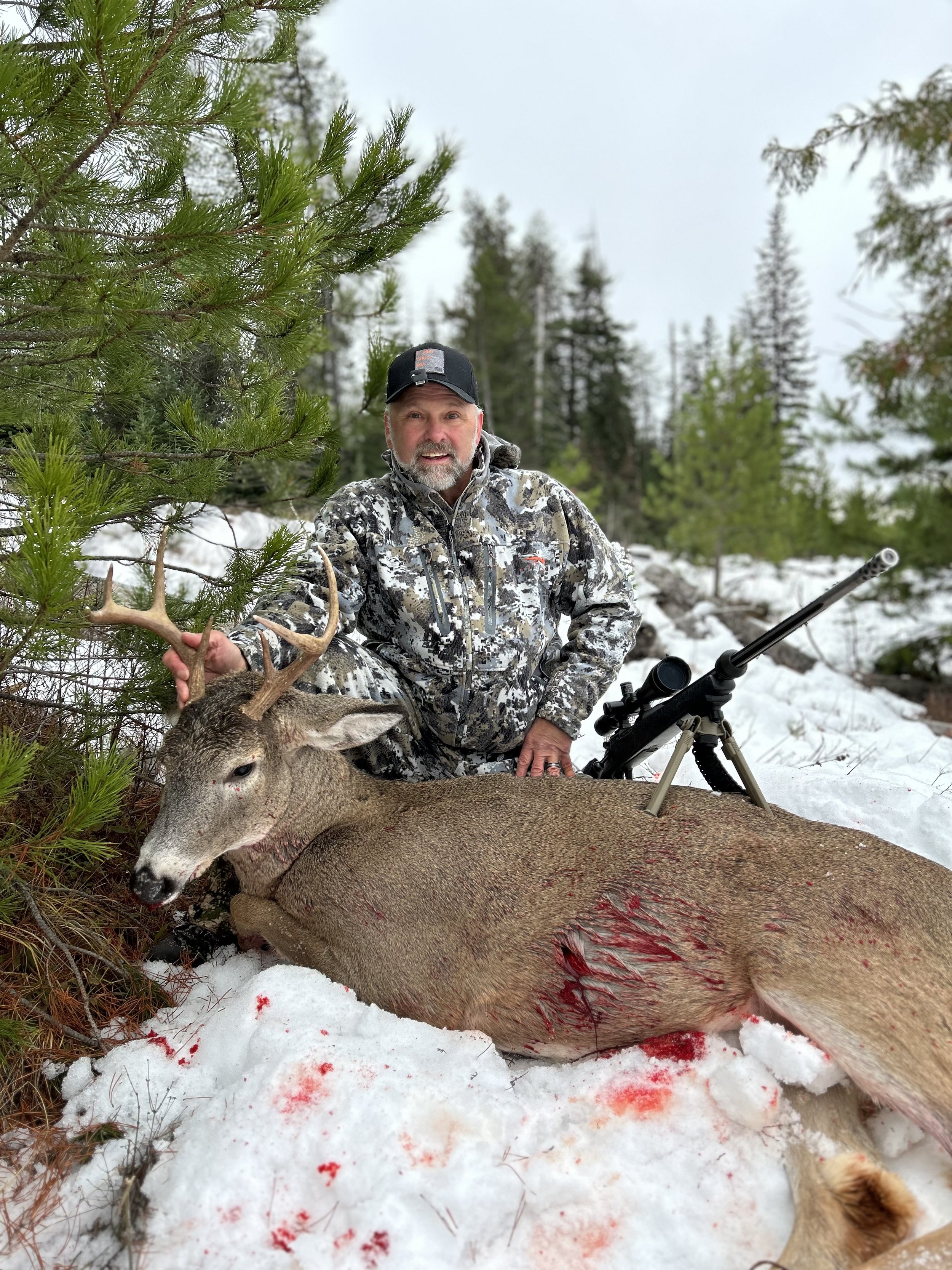 A man in camouflage clothing kneeling in the snow next to a large deer with antlers, a bloody wound, and a rifle beside him. The background features pine trees and a cloudy sky.