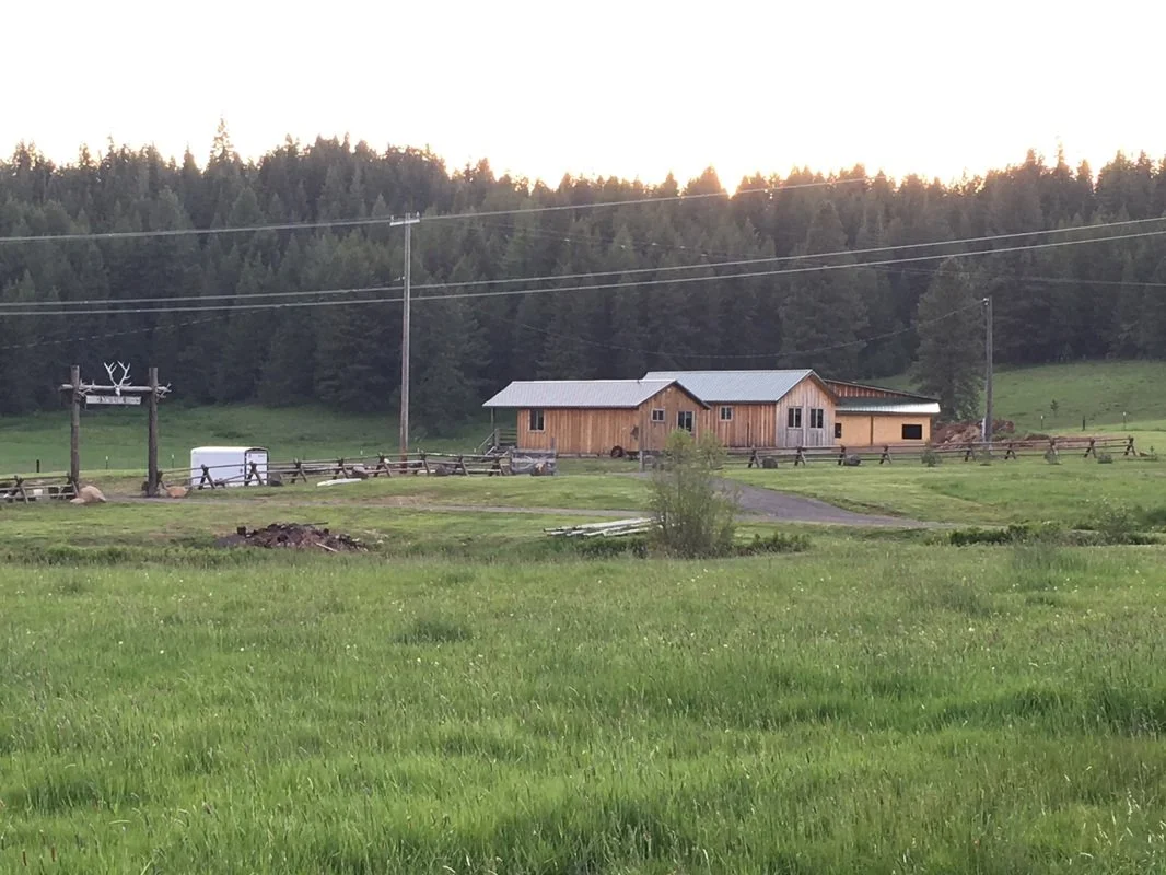 A rural farm scene with a wooden barn, green field, wooden fences, and a backdrop of forested hills under a cloudy sky at sunset.