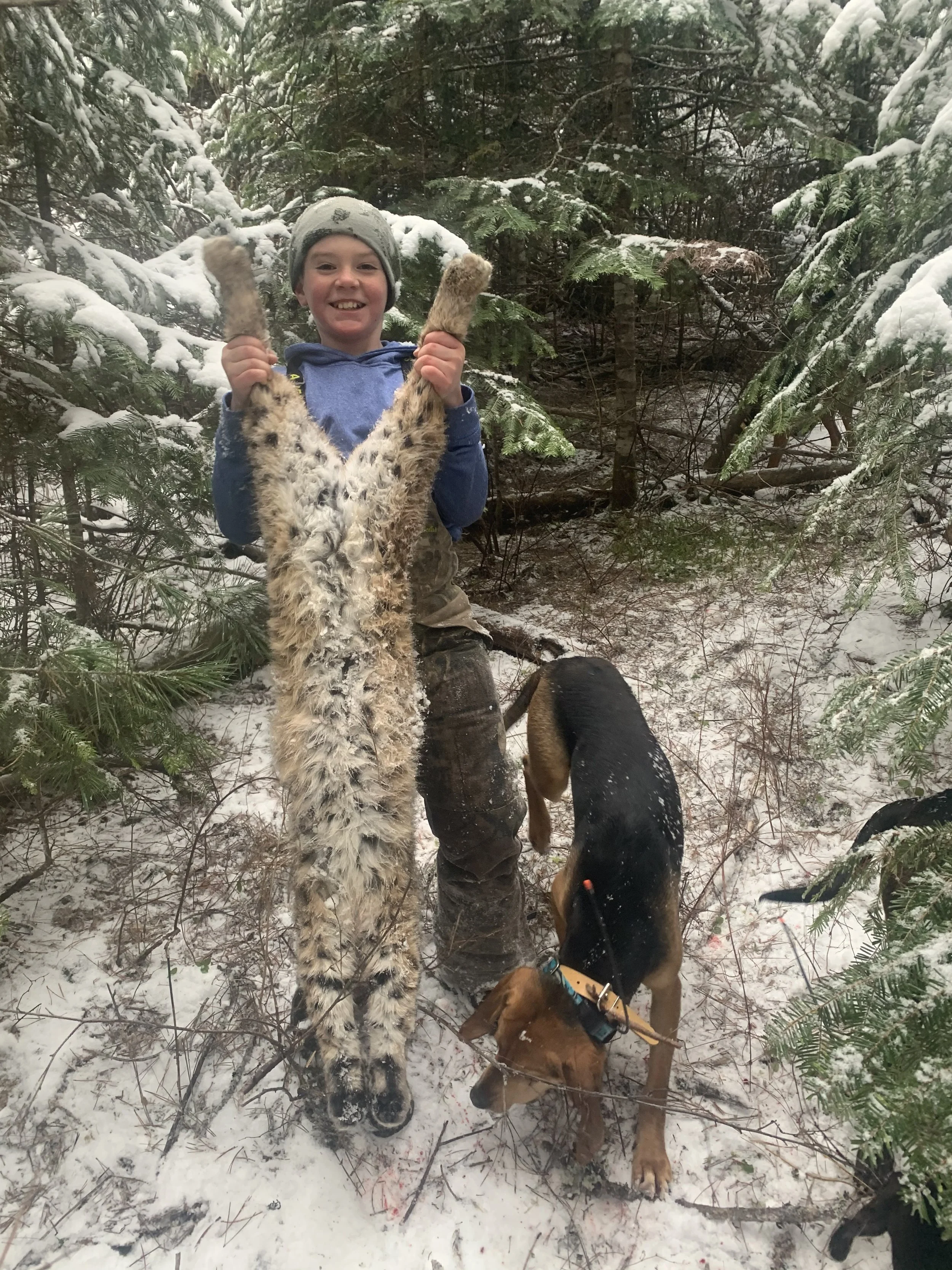 A young boy in a winter jacket and hat holding a caught bobcat upside down in a snowy forest, with a dog sniffing the ground nearby.