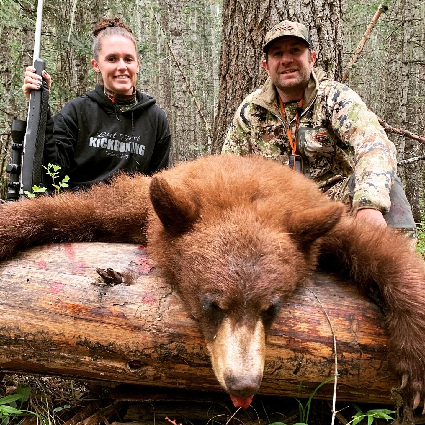 A man and woman in camouflage and black clothing with a rifle pose behind a large, bear lying on a fallen log in a forest.