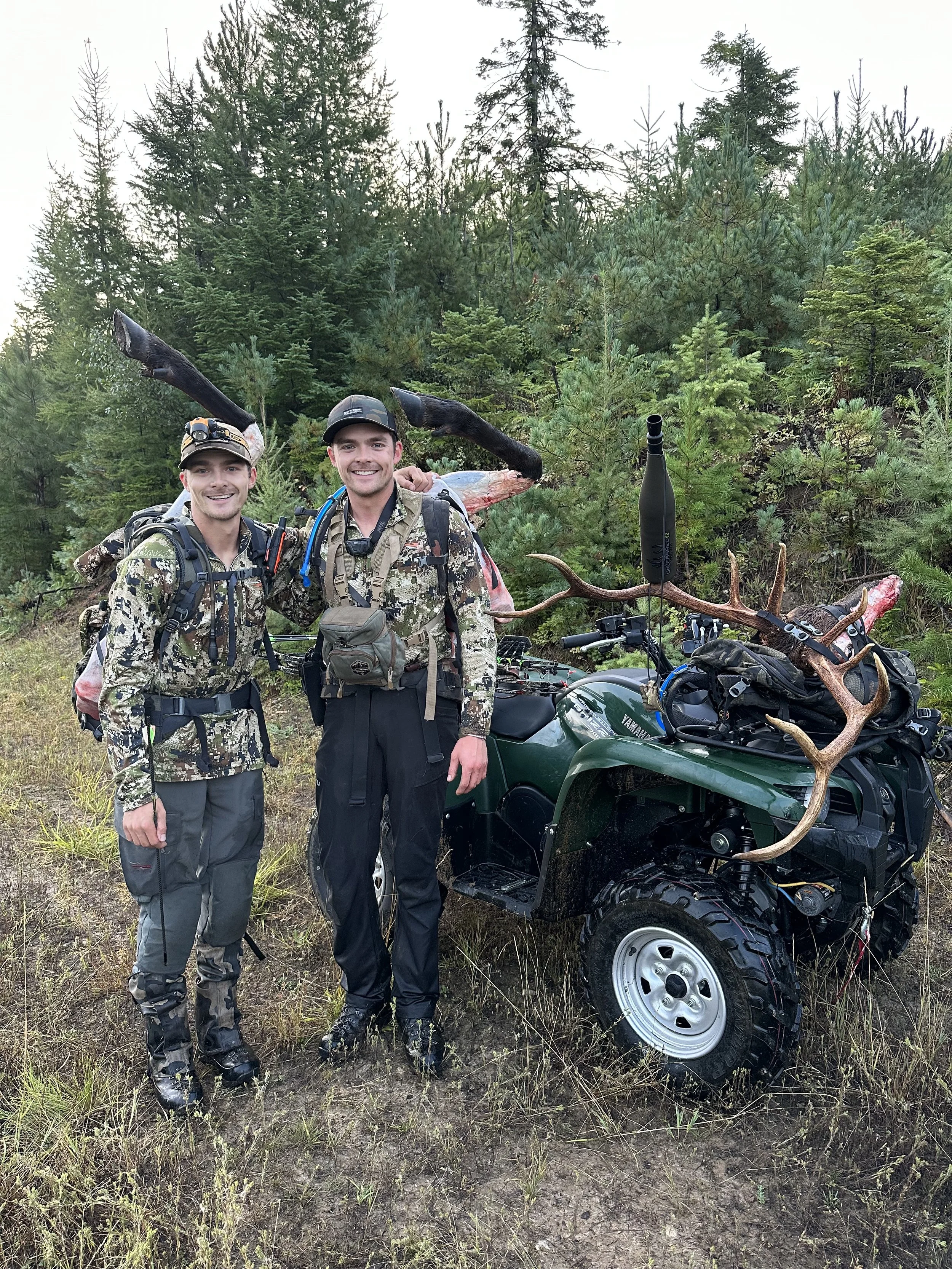 Two hunters in camouflage clothing standing next to an ATV with antlers and hunting gear, smiling after a successful hunt in a forested area.