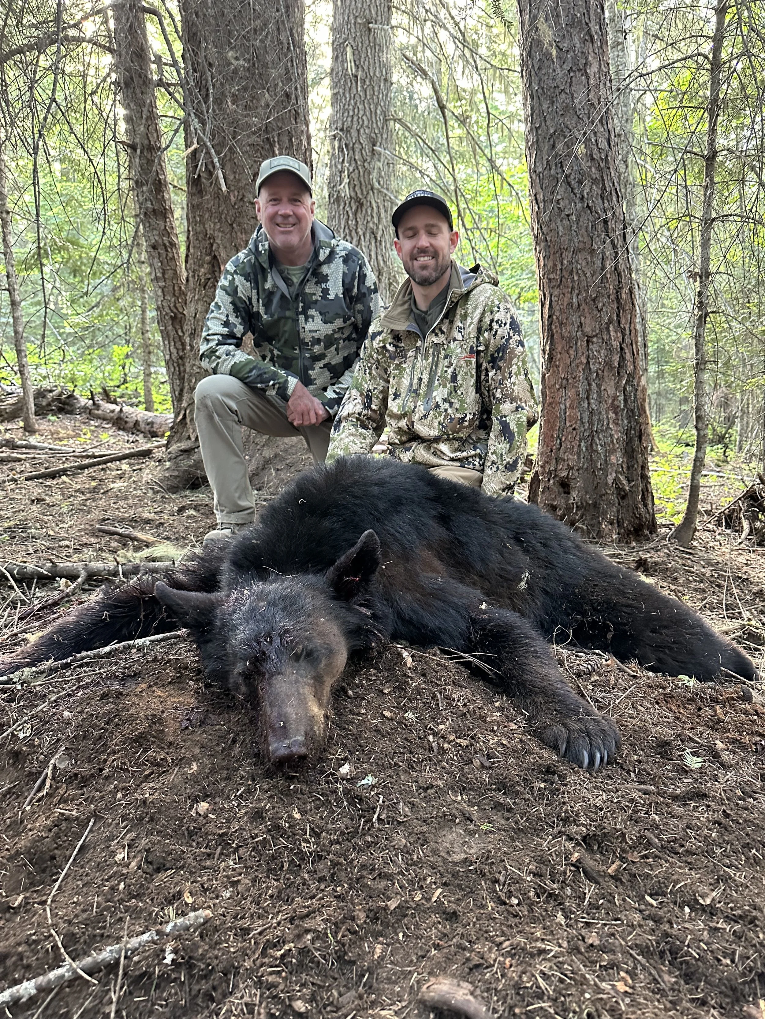 Two men in camouflage hunting outfits kneel behind a large, dead black bear lying on the ground in a forest with tall trees and green foliage.