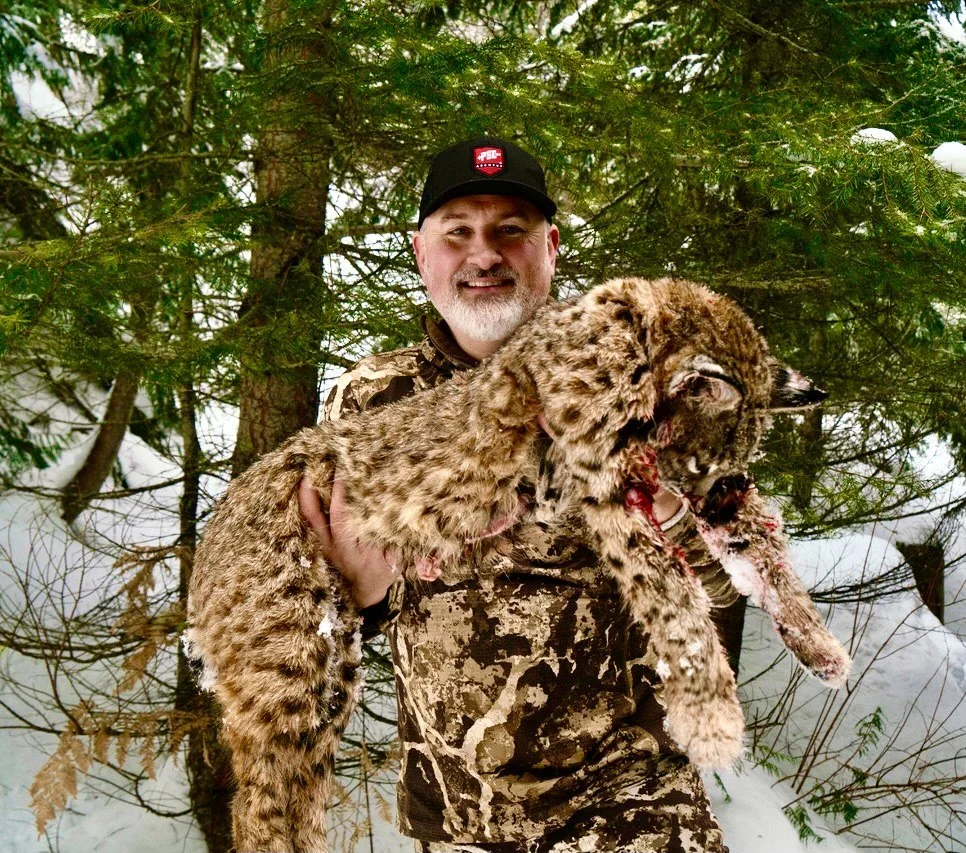 Man in camouflage jacket and cap holding a dead bobcat in a snowy forest.