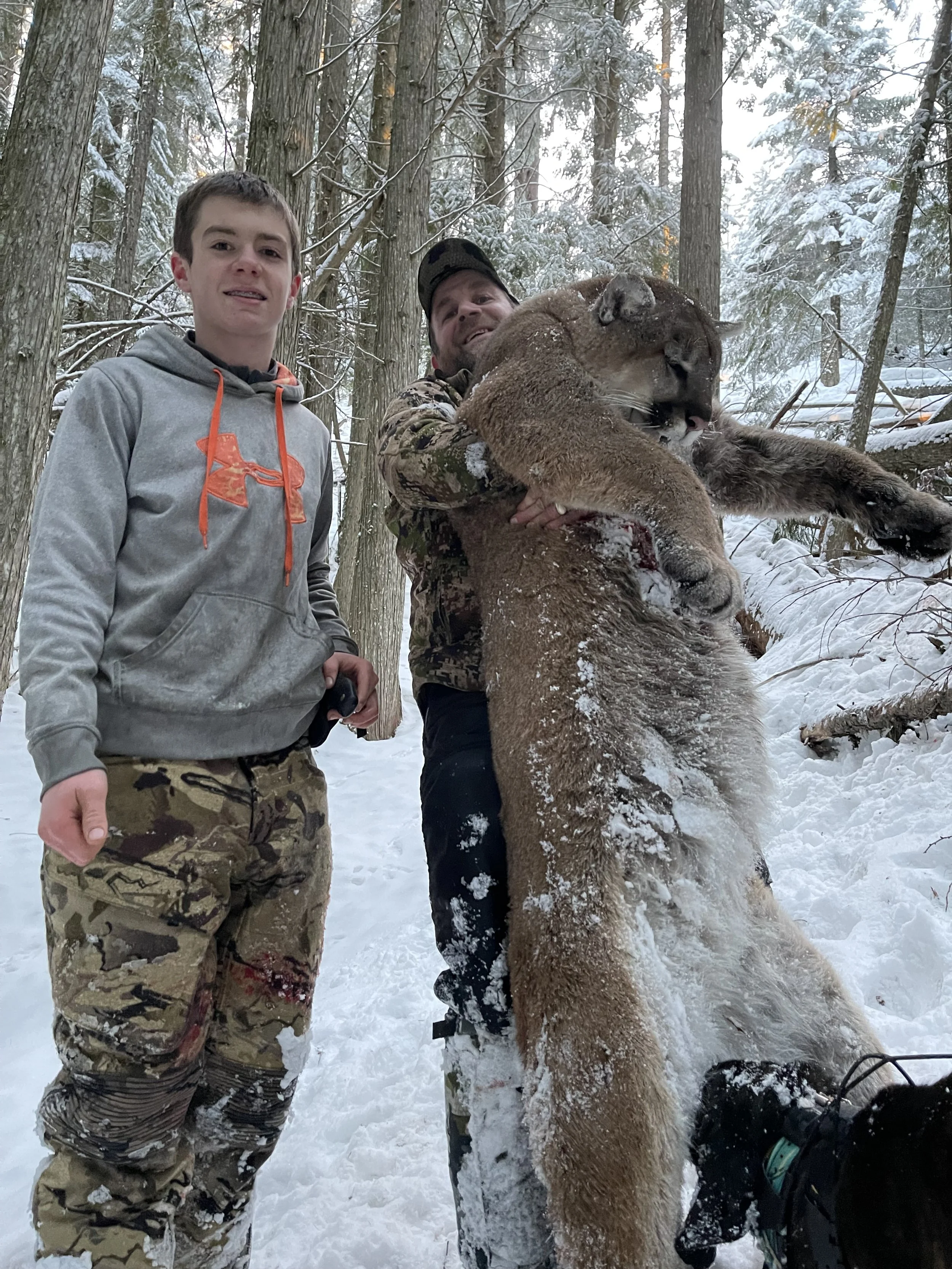 Two men and one young boy in a snowy wooded area, with one man holding a large, dead mountain lion and a puppy beside them.