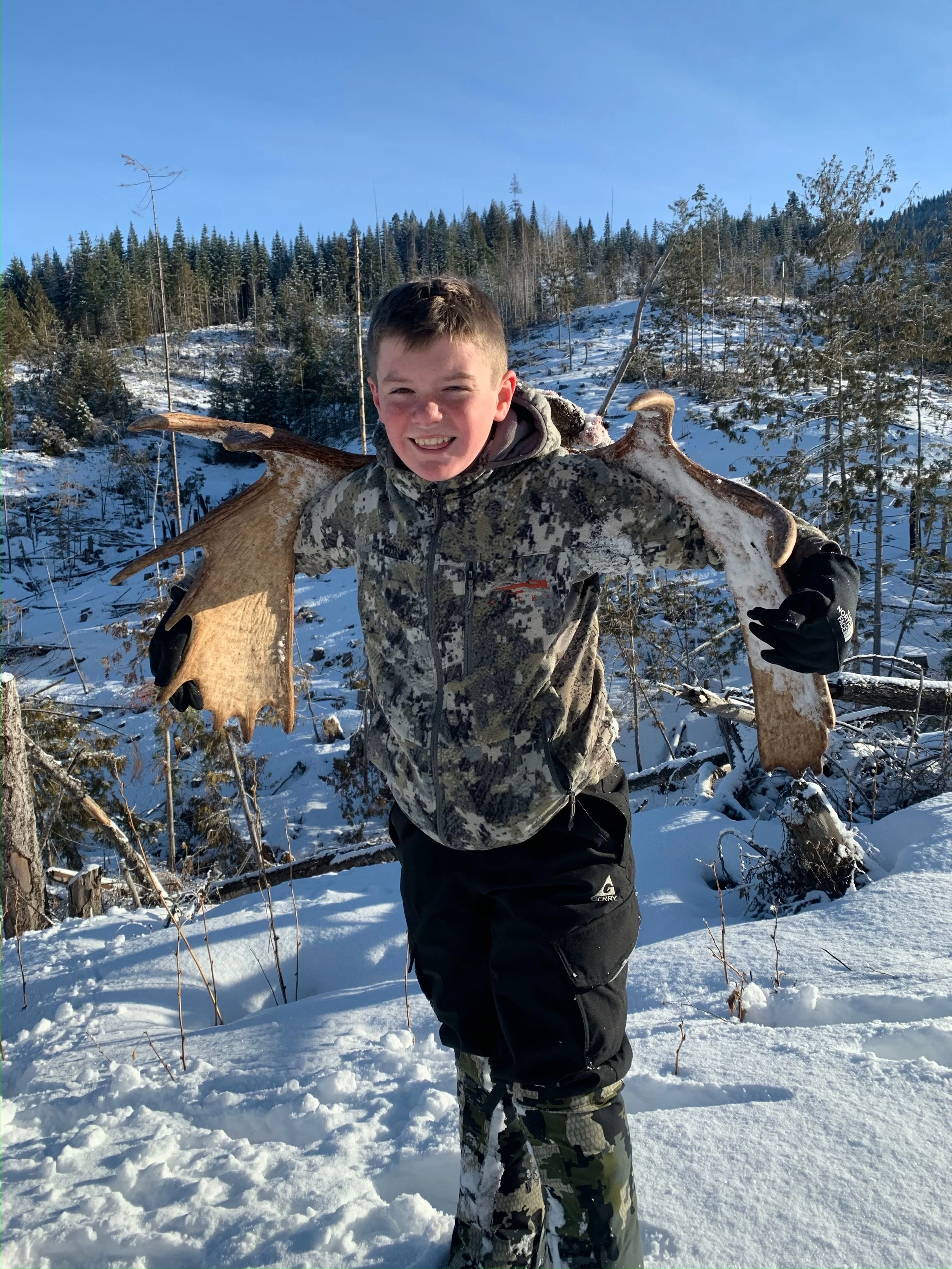 A boy standing in snowy woods holding large moose antlers across his shoulders, smiling at the camera.