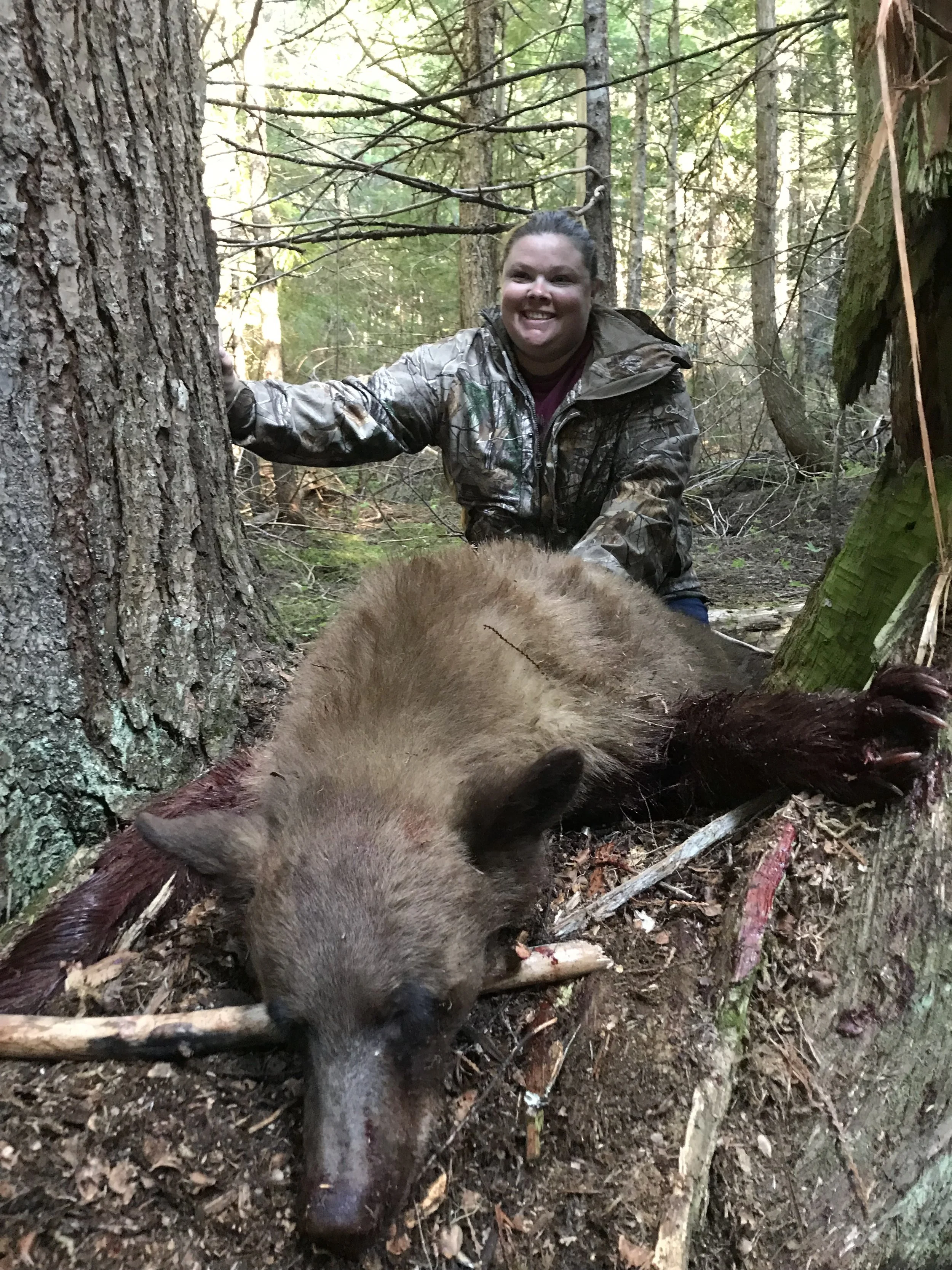 A woman in camouflage clothing smiling in a forest next to a large dead wild boar lying on the ground with a stick through its mouth.