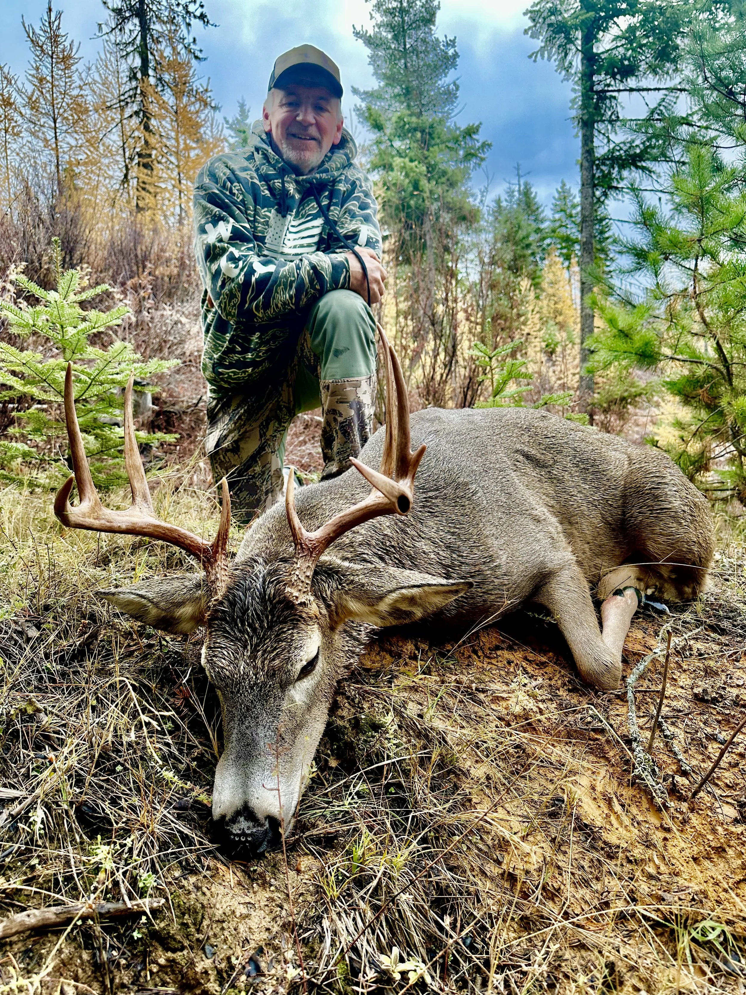 A man in camouflage clothing kneels beside a large, deceased deer with antlers in a forested area during daytime.