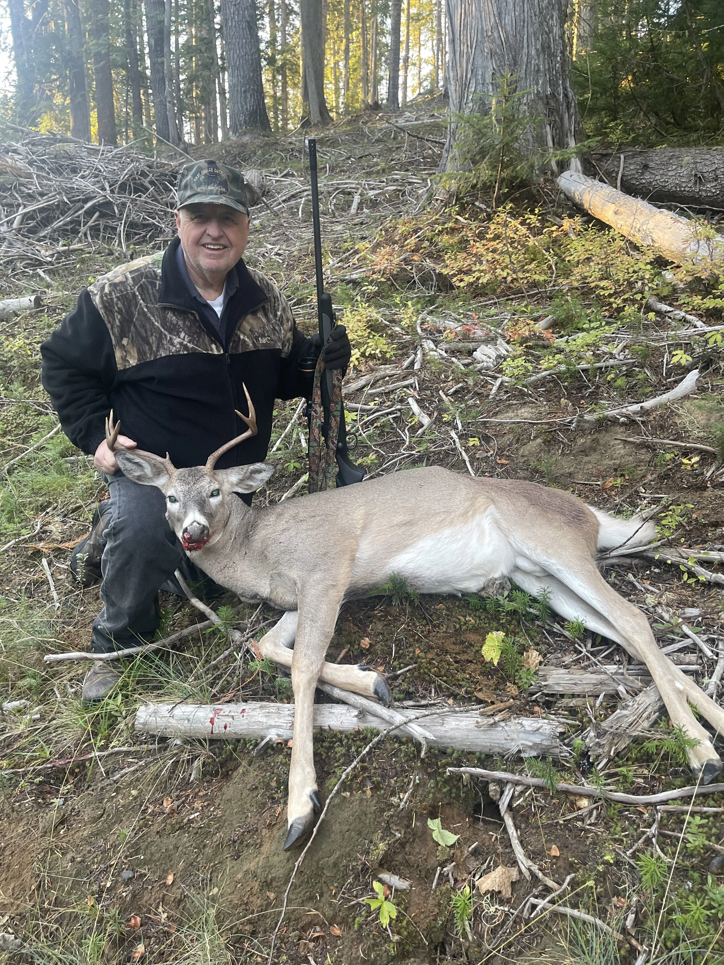 Man in camouflage and black hunting gear kneels next to a killed deer with antlers, holding a rifle, in a forested area with trees and fallen branches.