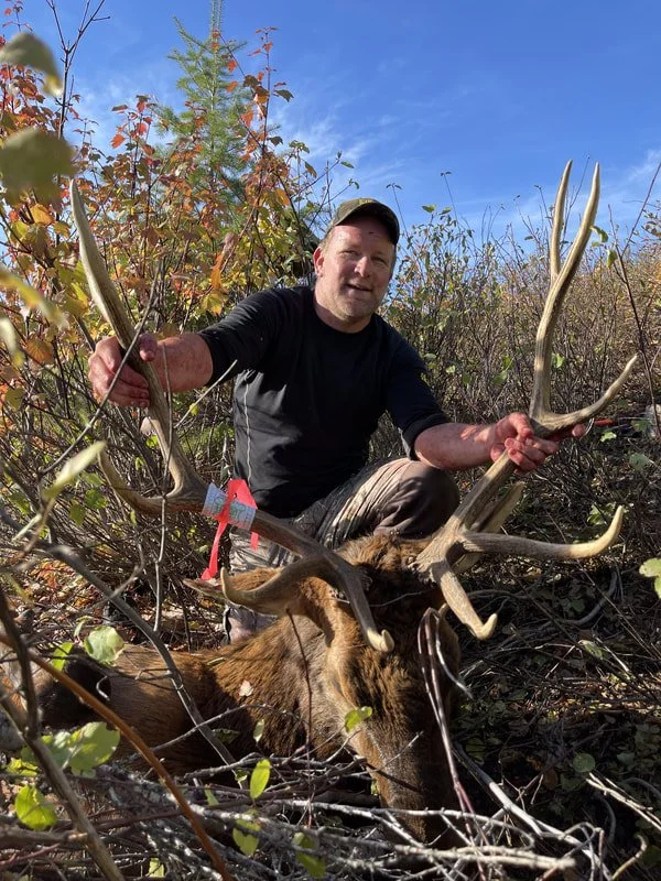A man outdoors holding a large deer with prominent antlers after a hunting trip, surrounded by bushes and trees under a blue sky.