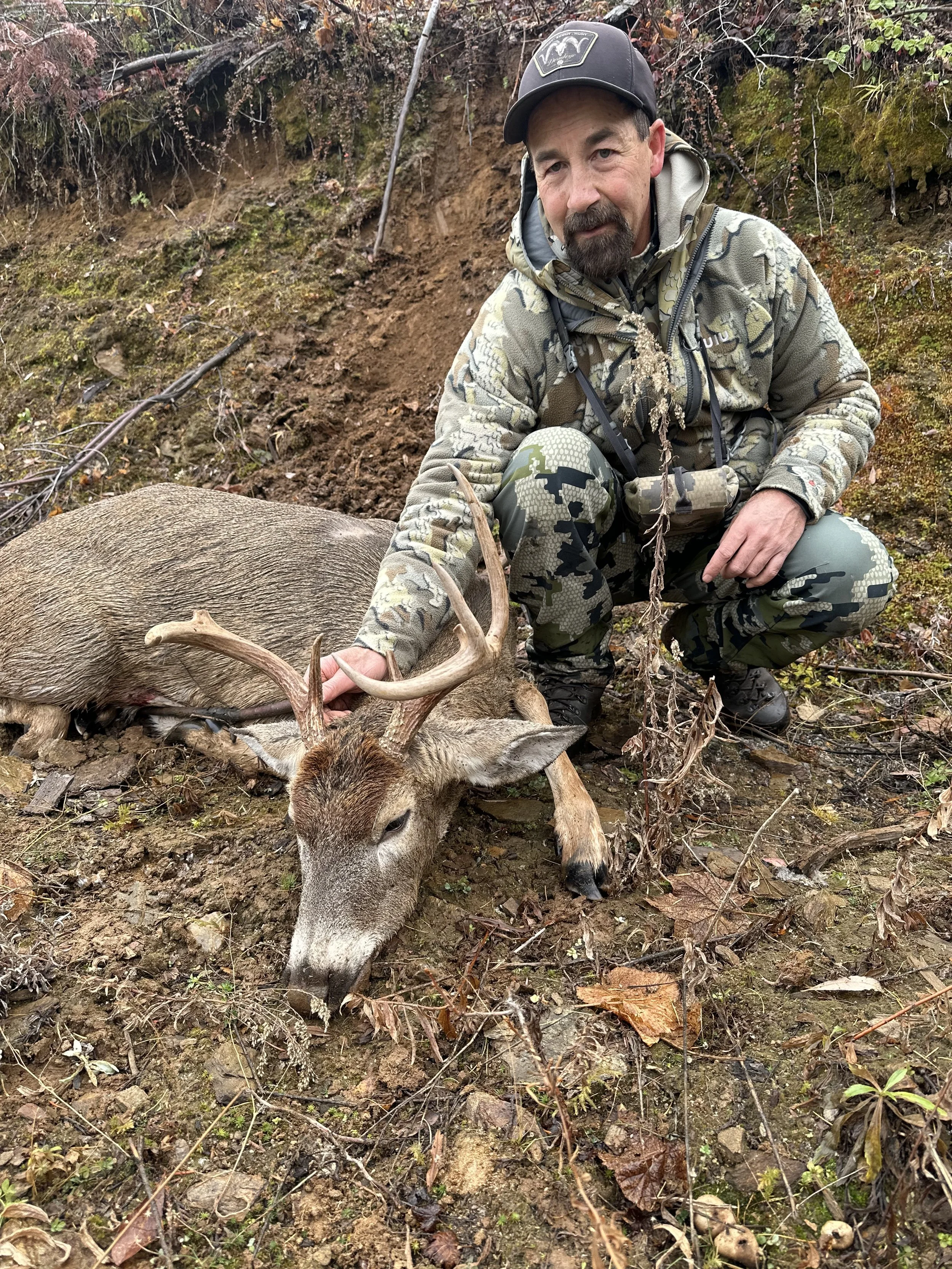 A man in camouflage clothing kneels next to a fallen deer with antlers in a muddy, wooded area.