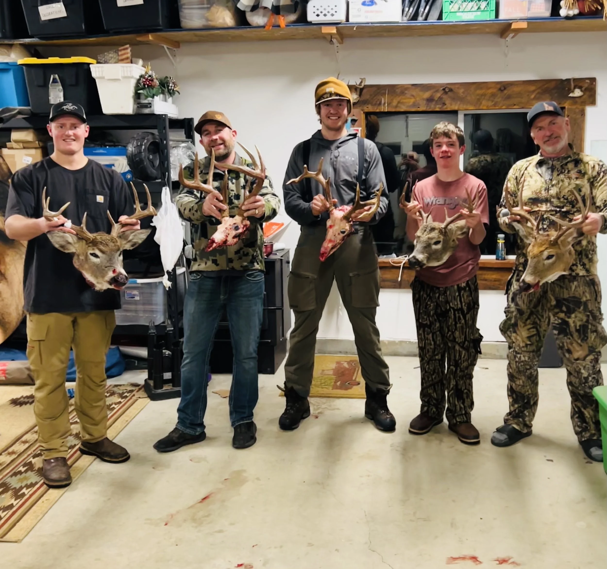 Five men standing indoors, each holding a deer head with antlers and the carcass, posing in a garage or storage room. The room has shelves with boxes and supplies in the background.