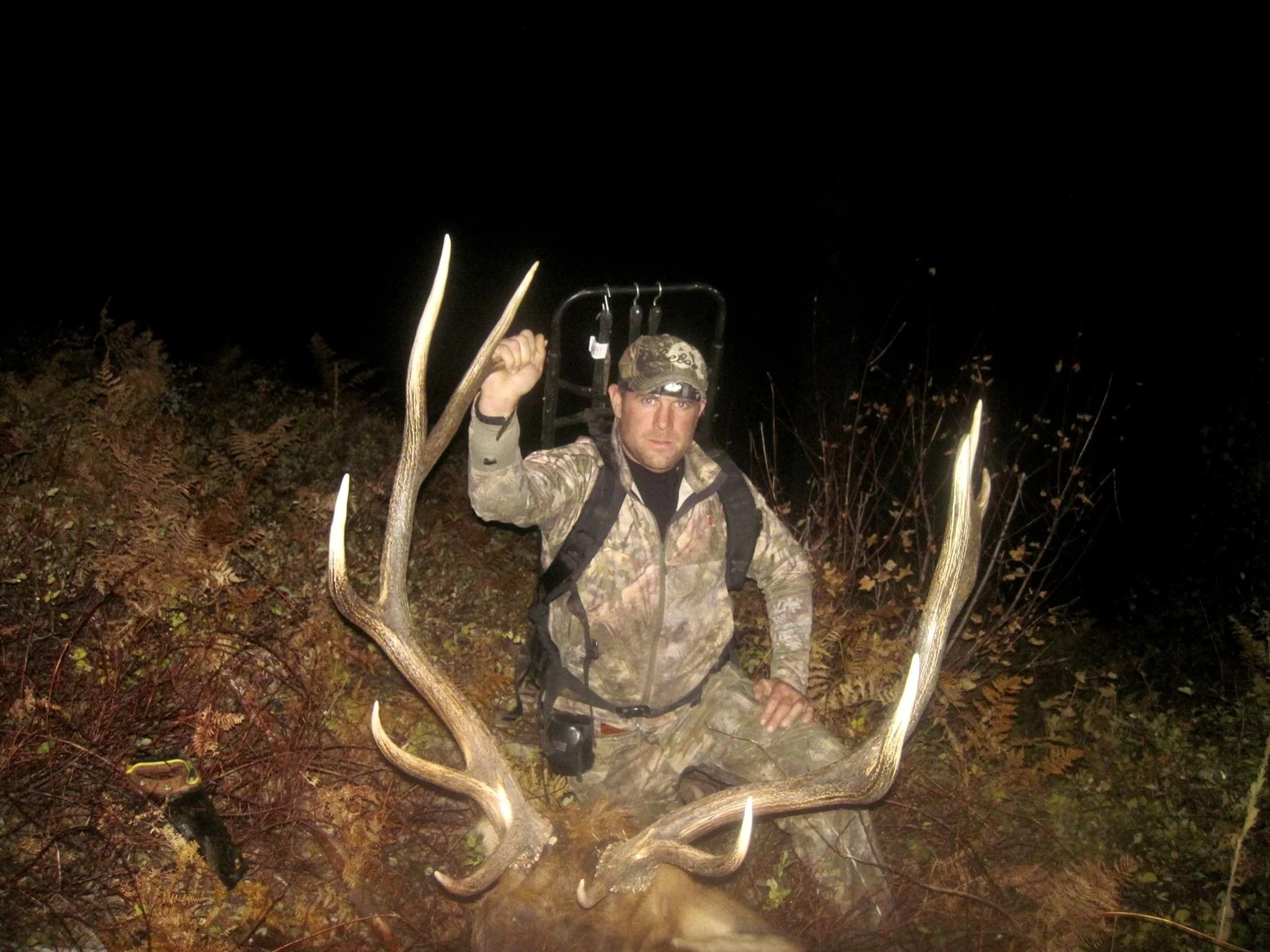 A man in camouflage clothing sitting with a large deer he hunted at night, holding its antlers visible in the foreground.