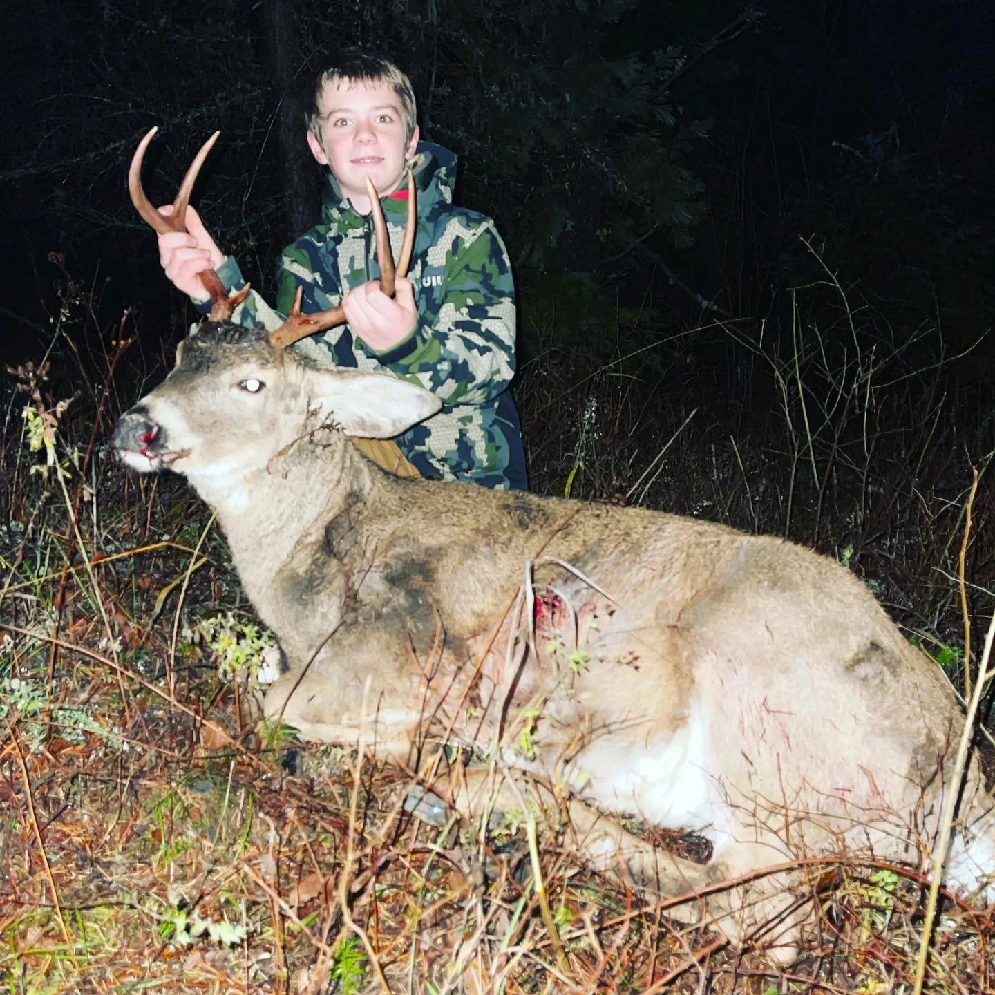 A boy in camouflage clothing holding the antlers of a large, recently hunted deer with a bleeding wound, standing in a dark wooded area at night.