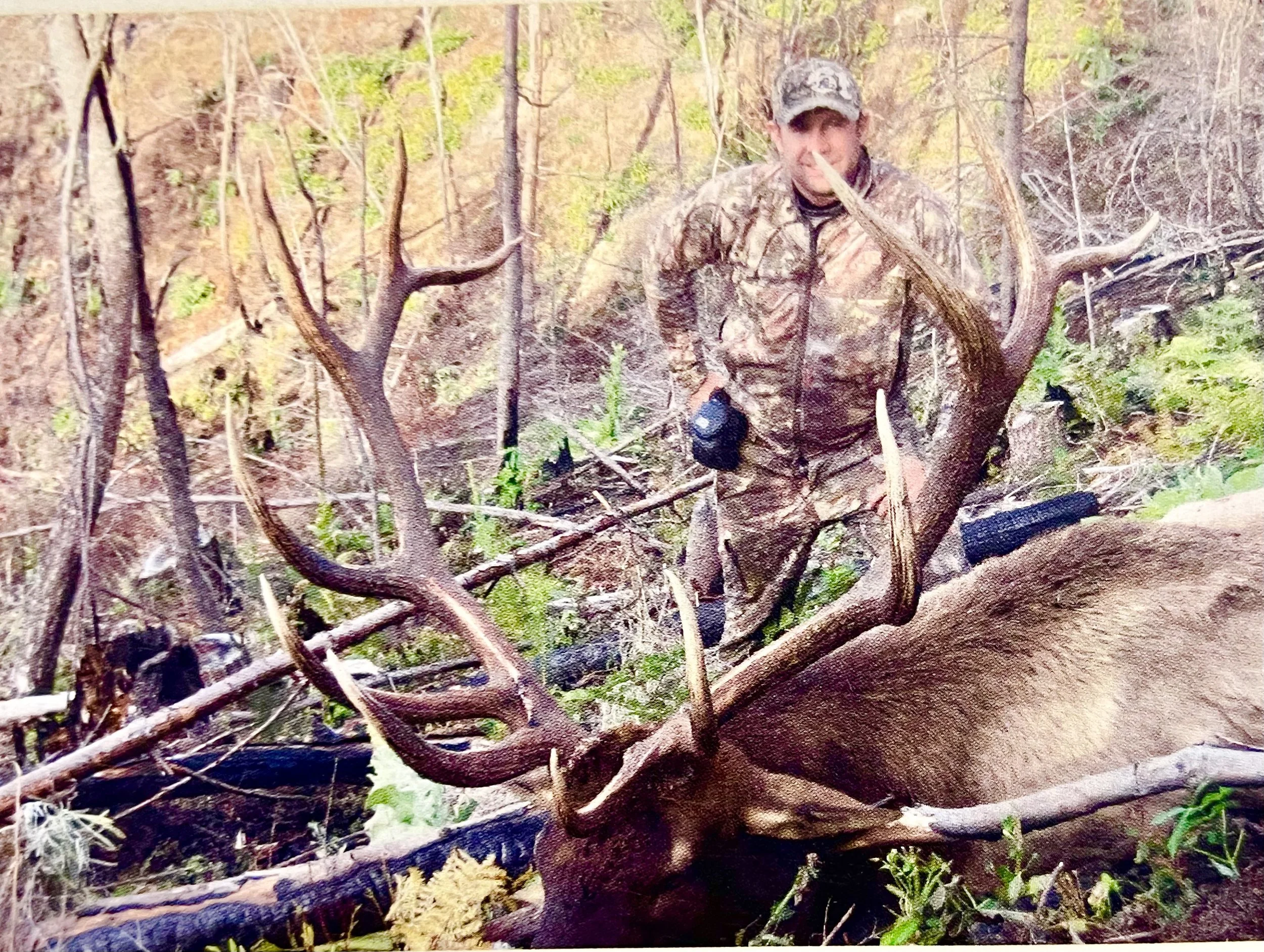 A man dressed in camouflage clothing standing in a forested area next to a large dead elk with impressive antlers, surrounded by fallen branches and trees.