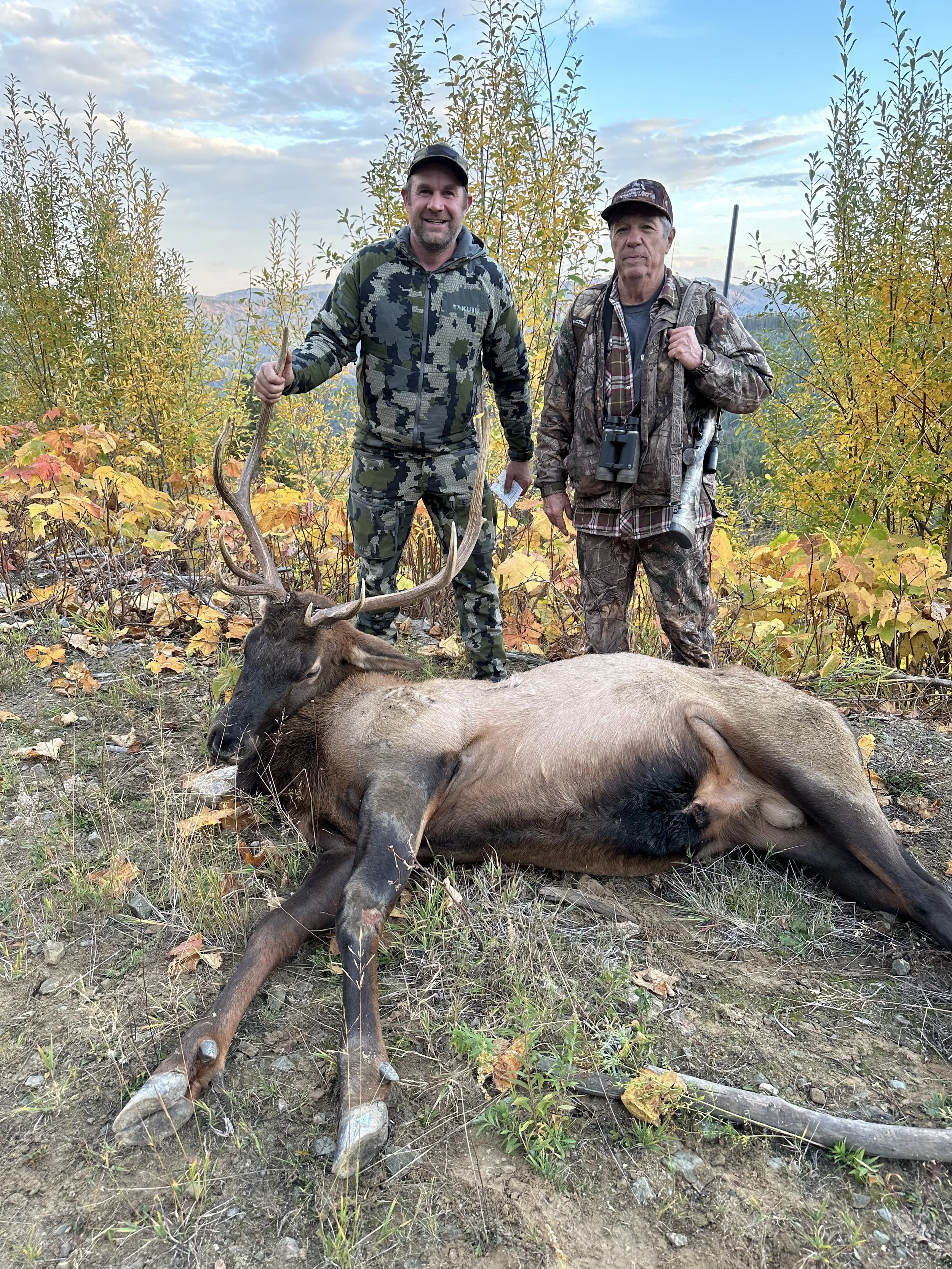 Two hunters in camouflage clothing stand behind a deer with antlers that they have hunted, in an autumnal forest with yellow and green leaves, under a partly cloudy sky.