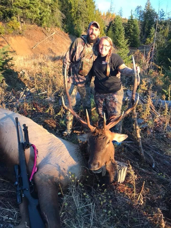 Two people, a man and a woman, stand outdoors in a forested area, posing next to a large elk they have hunted. The woman holds the elk's antlers, and a rifle rests on the elk's body.